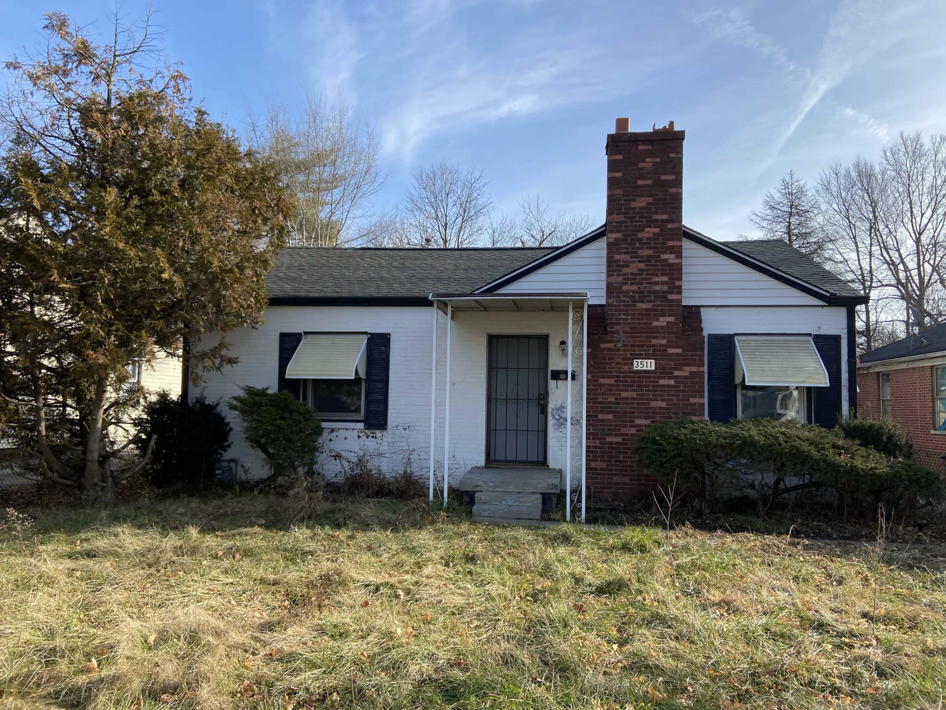 A single-story house with a brick chimney and overgrown yard, featuring white siding and dark blue shutters.