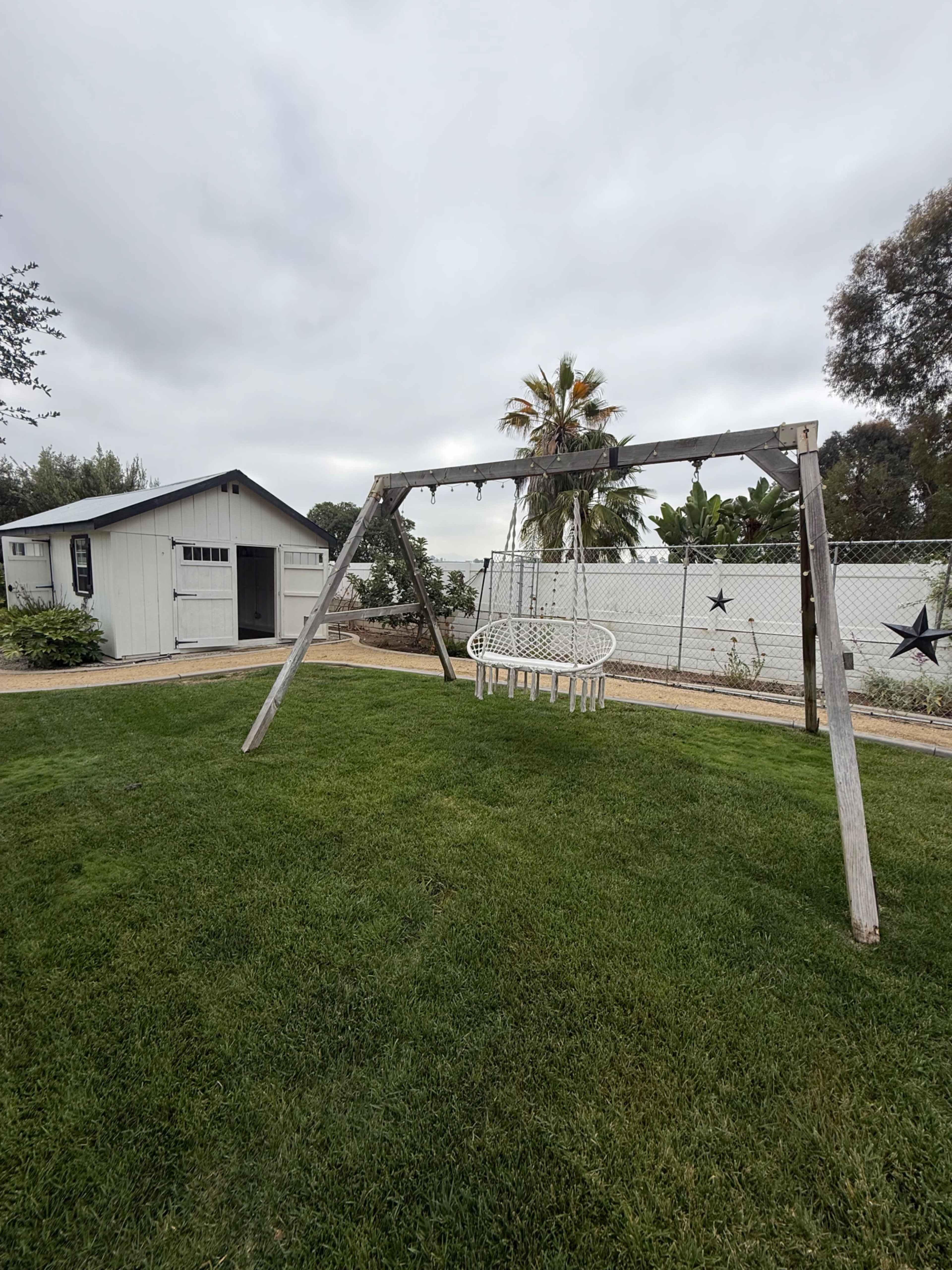 A wooden swing set with a hanging chair is placed on a grassy lawn next to a shed and surrounded by palm trees and a fence.