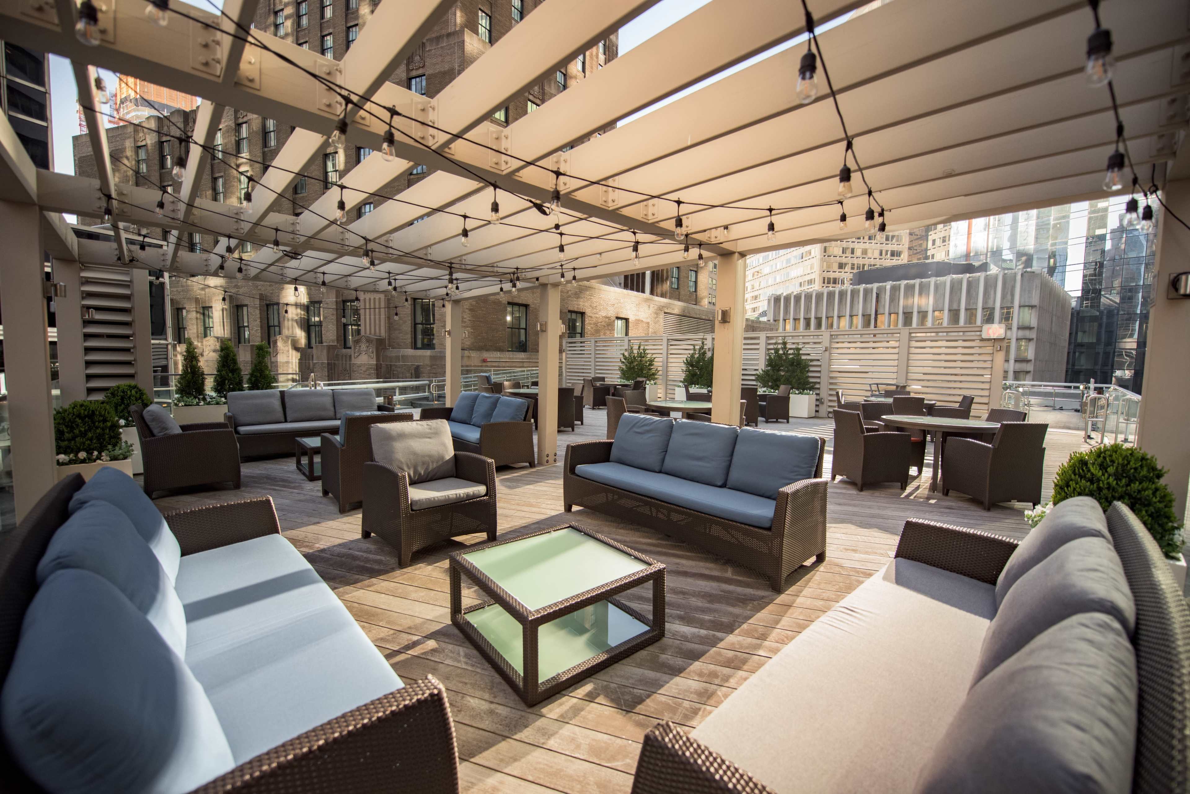 A rooftop patio with several seating areas, including sofas and tables, illuminated by string lights against a backdrop of urban buildings.