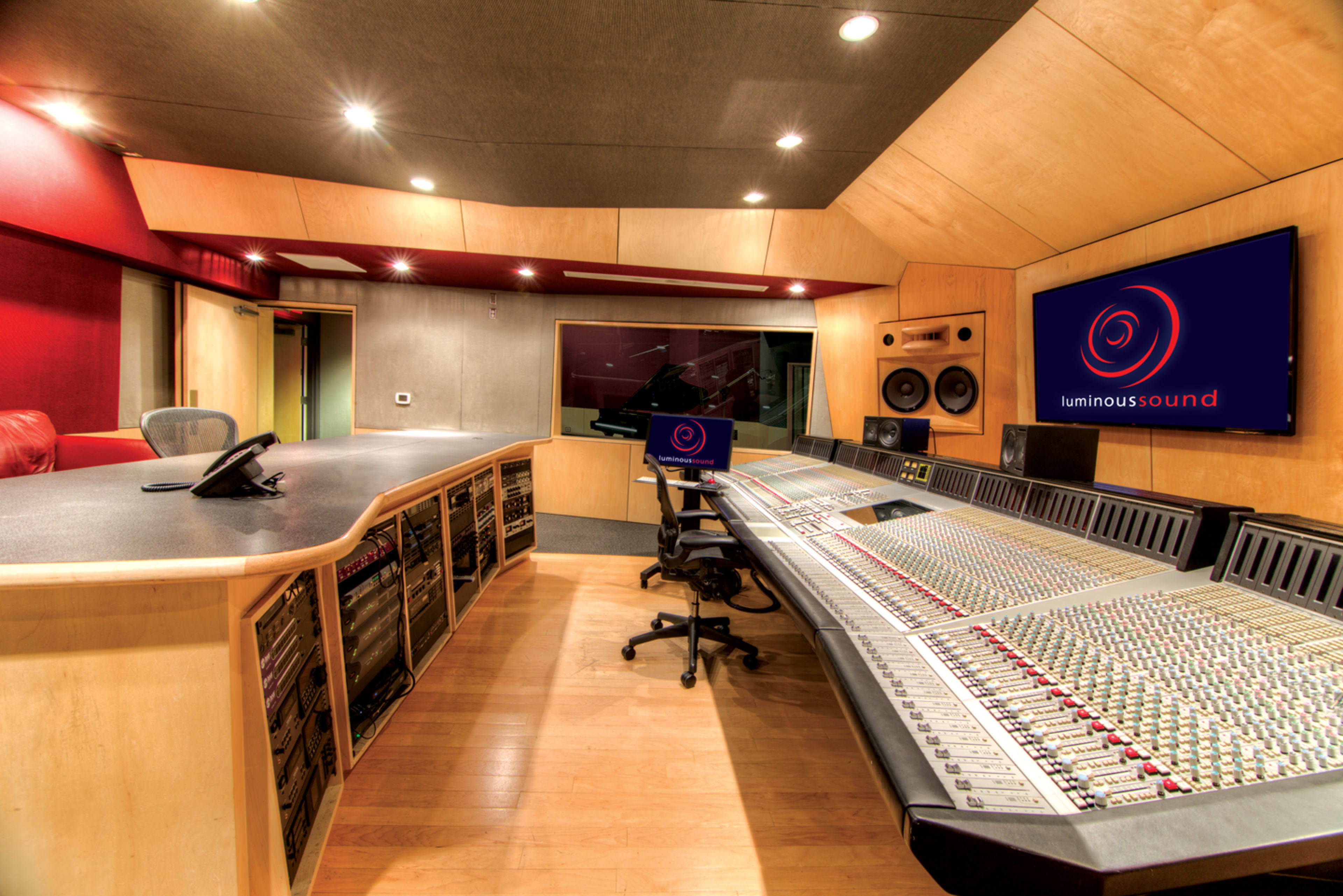 The image shows a professional audio recording studio featuring a large mixing console, control desk, and wall-mounted screens.