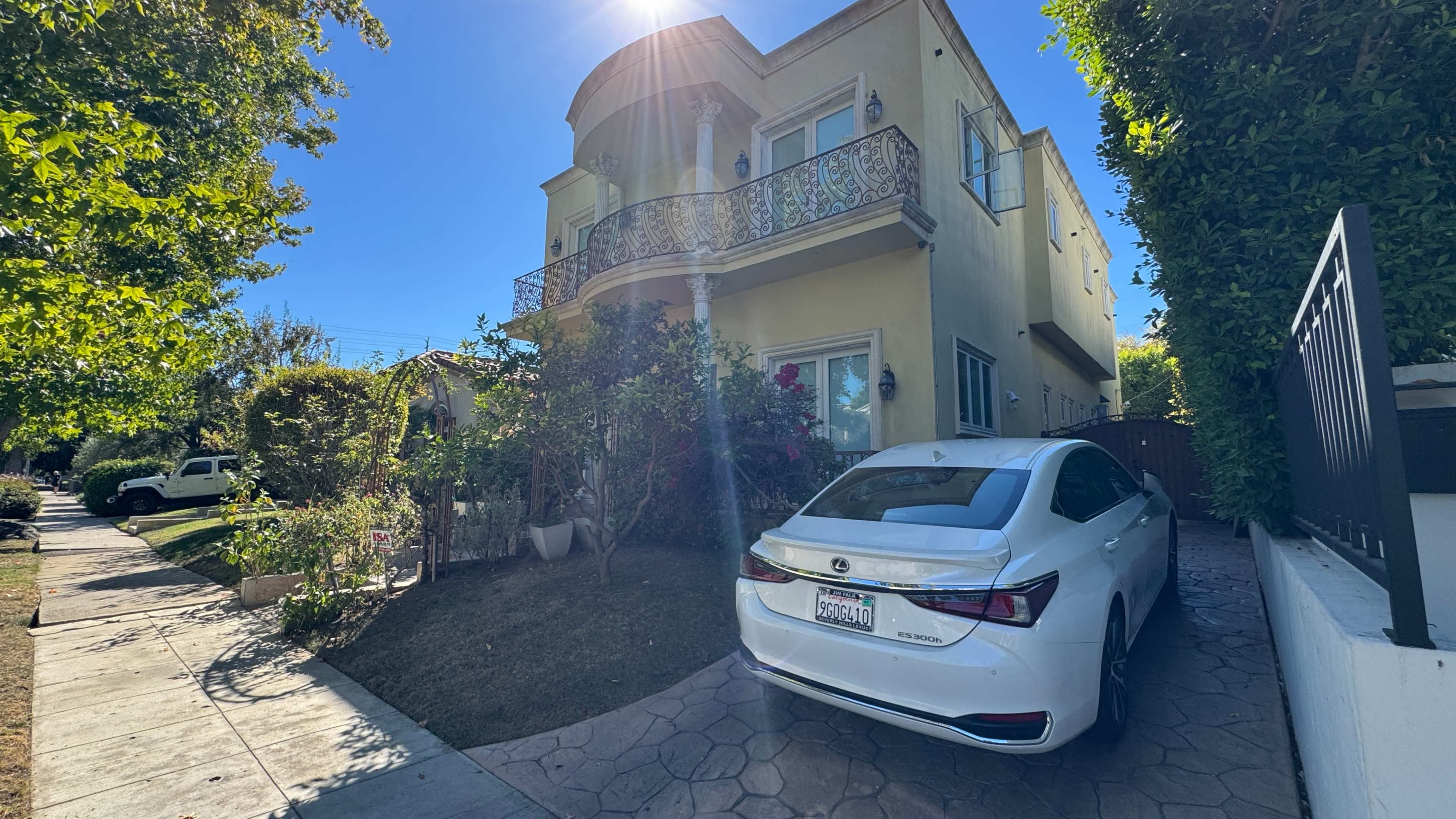 A white car is parked in front of a two-story yellow house with a balcony, surrounded by greenery and flowers.