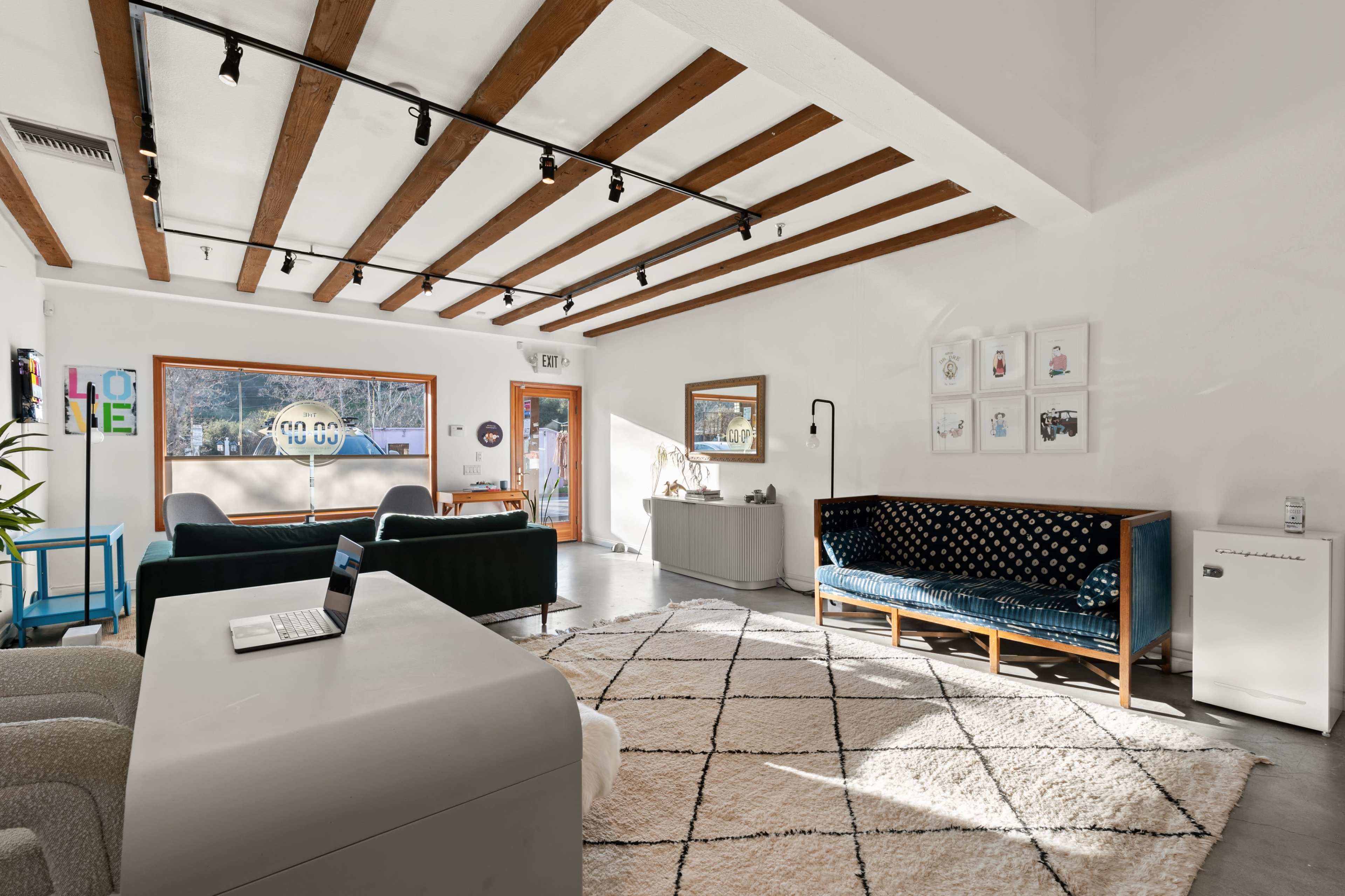 A modern living space featuring exposed wooden beams, a rug on a concrete floor, and a mix of contemporary furniture including a blue couch and a grey sideboard.