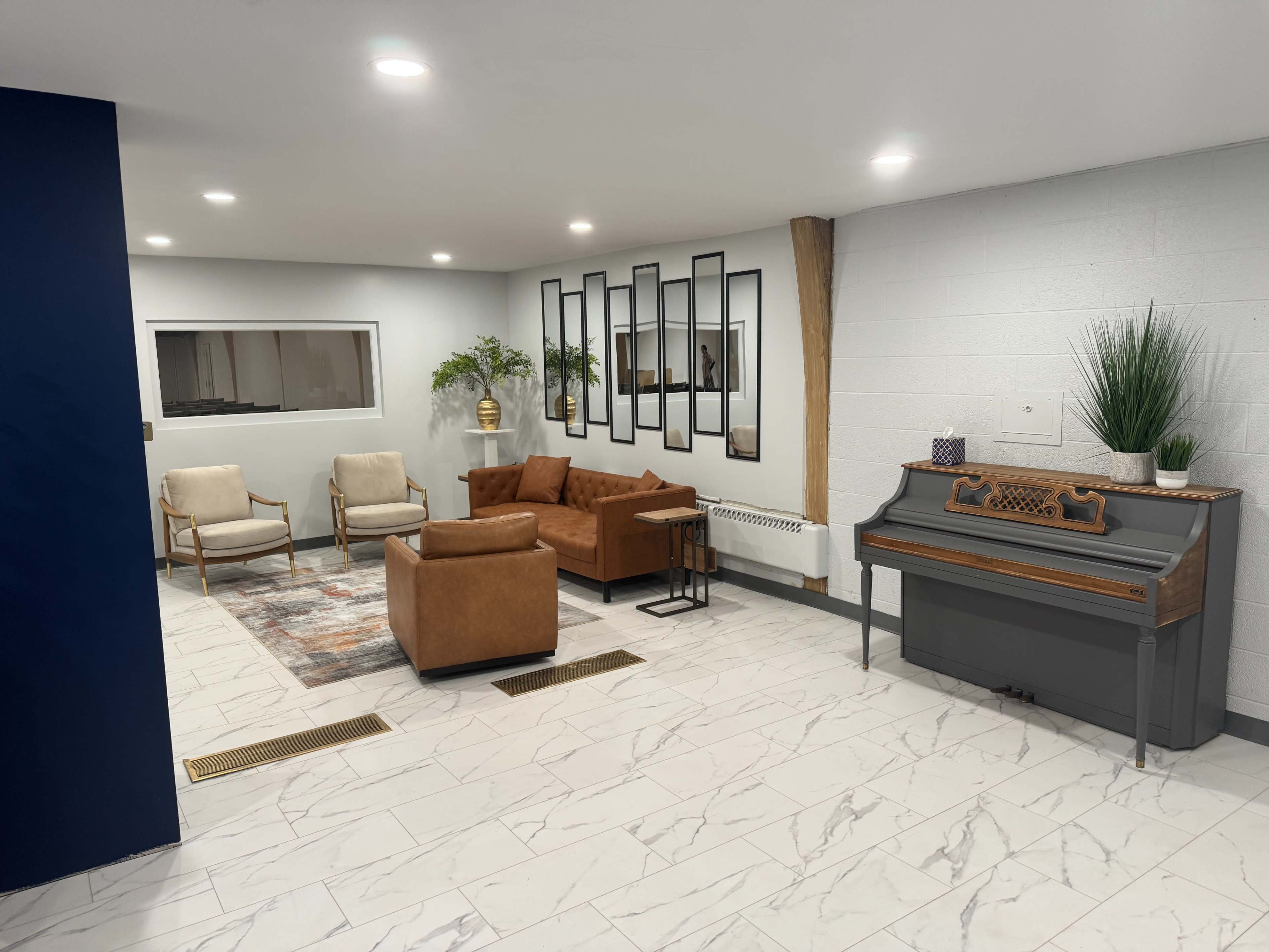 The image shows a modern, well-lit interior space featuring a brown leather sofa, two light-colored chairs, a decorative mirror arrangement on the wall, and a piano in the corner.