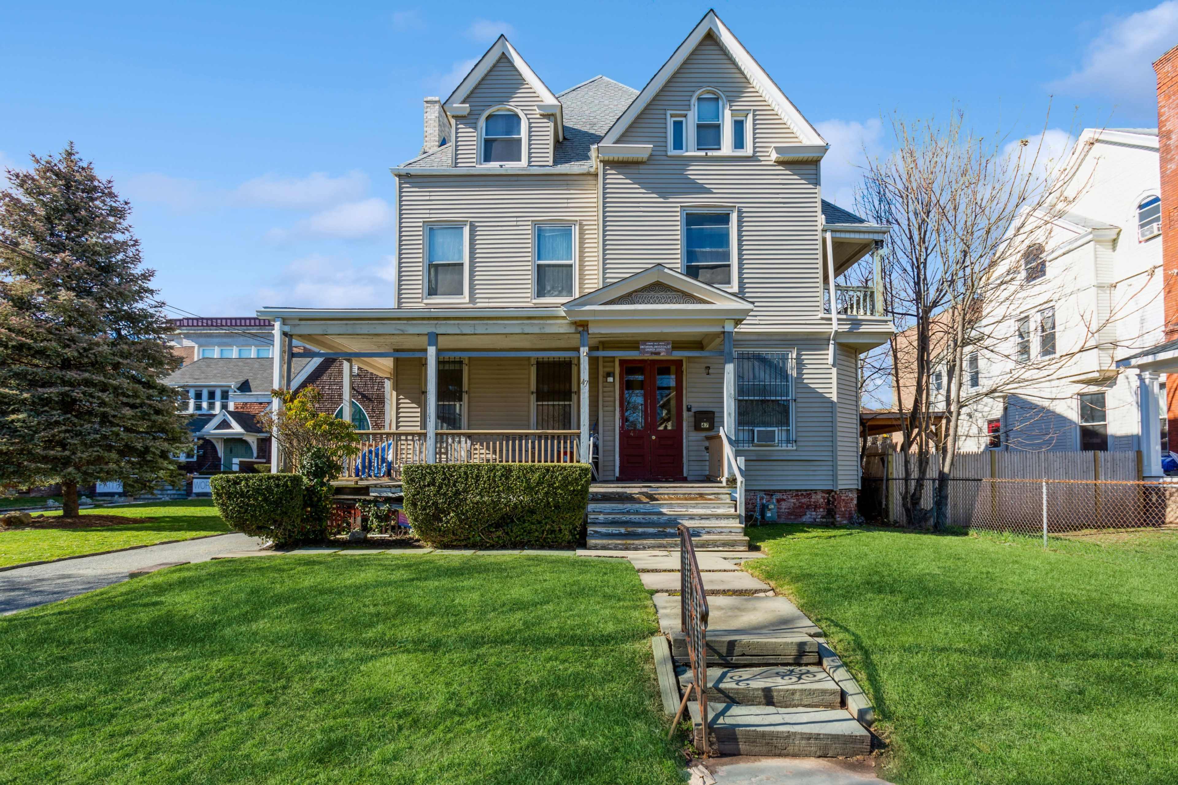 A large, multi-story house with a front porch and a manicured lawn is shown against a clear blue sky.