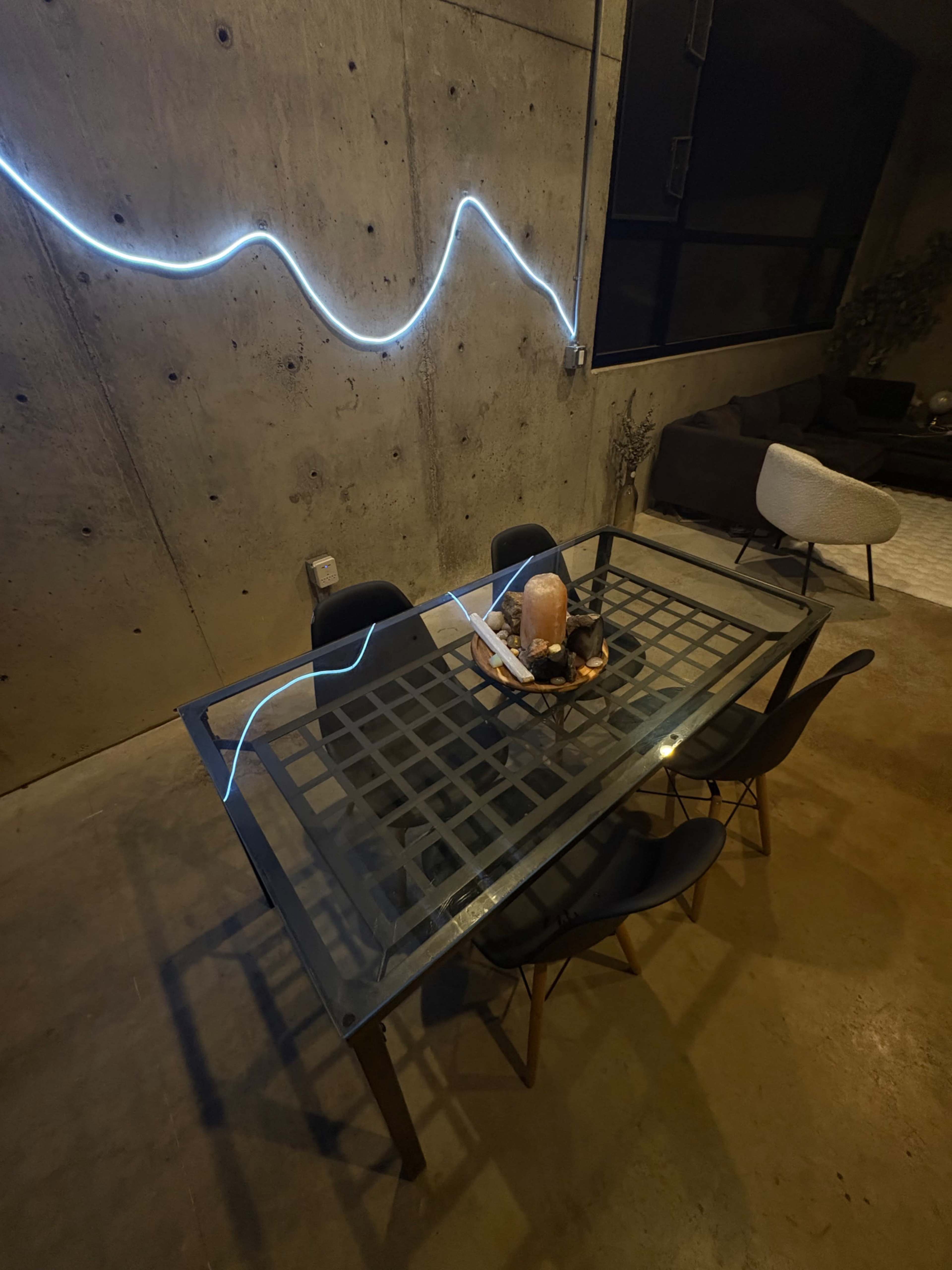 A glass-top dining table with black chairs is positioned on a concrete floor, illuminated by a blue neon light along the wall.