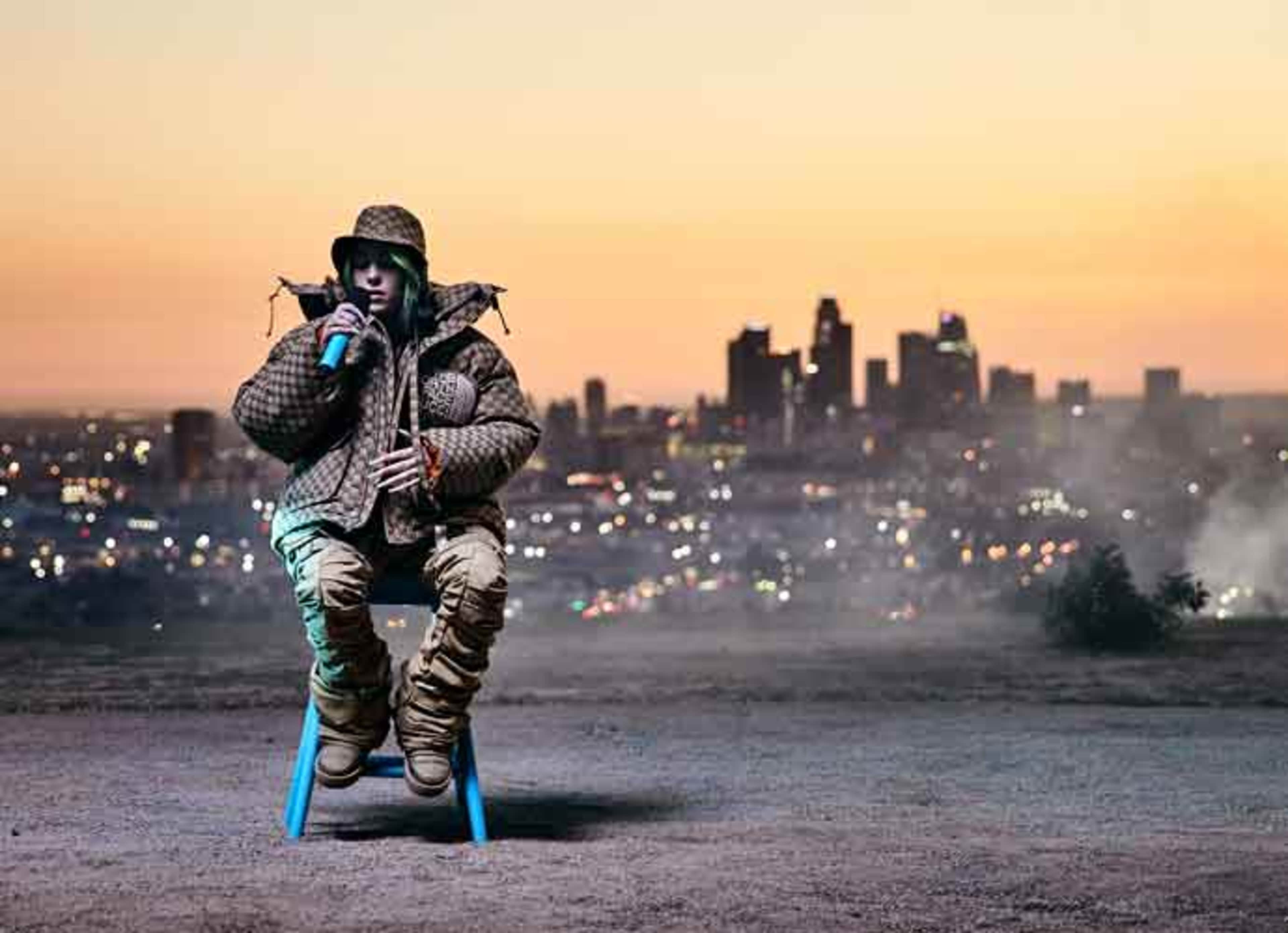 A person wearing a patterned coat and khaki pants sits on a blue chair against a city skyline at sunset.