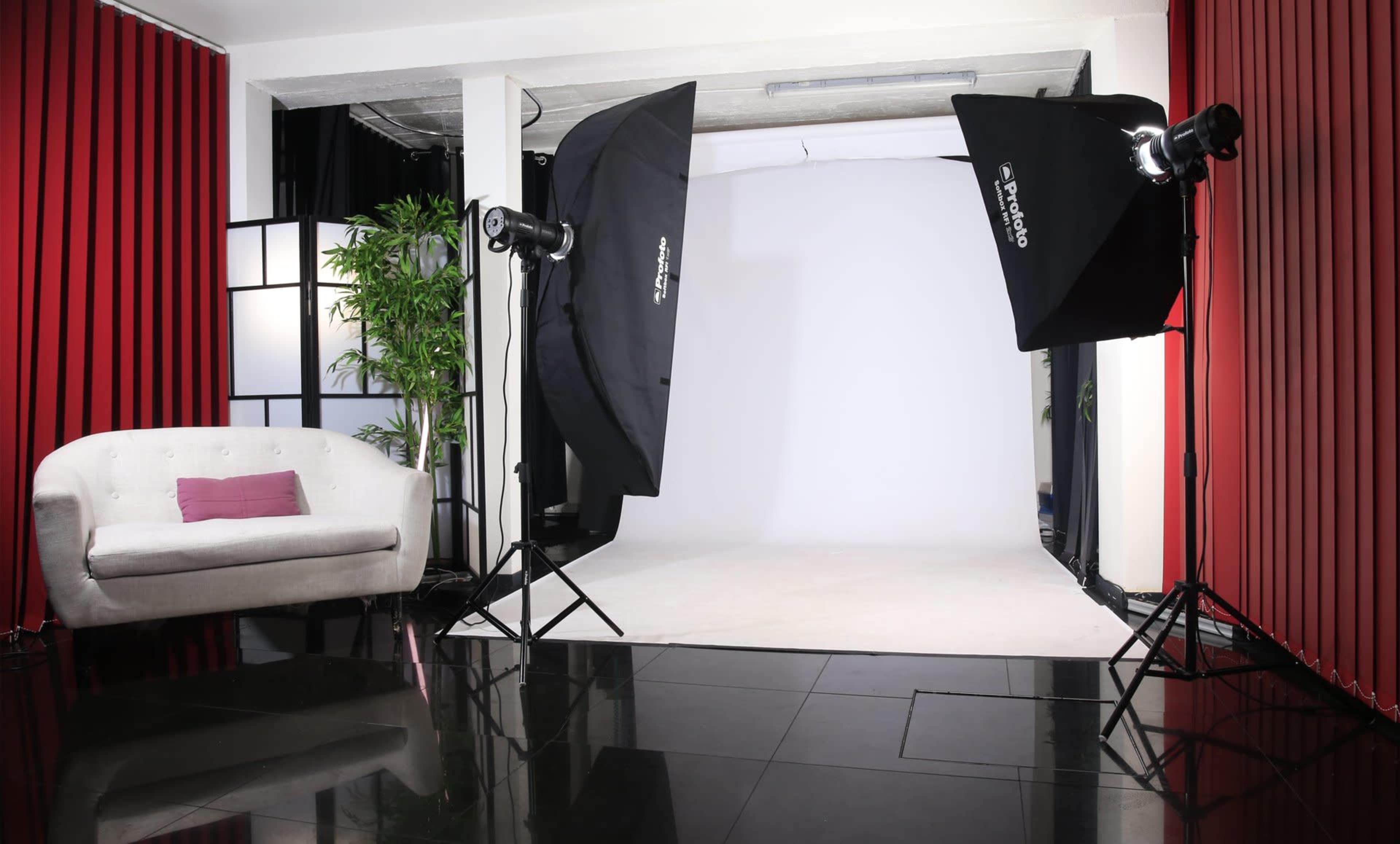 The image shows a photography studio setup with a white backdrop, two softbox lights, a black and white screen, a potted plant, and a white sofa.