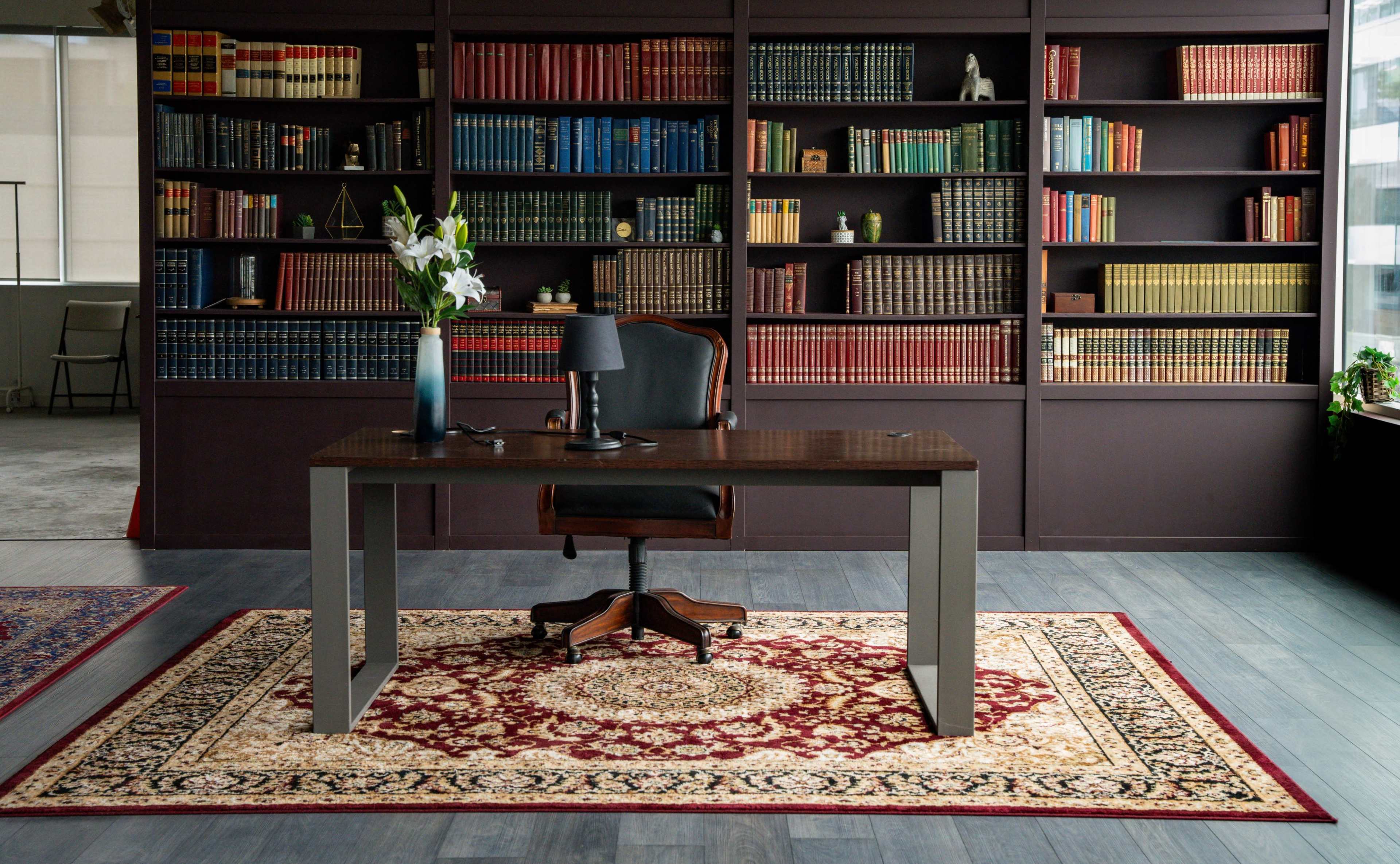 A wooden desk with a black chair is positioned on a patterned rug in front of a bookshelf filled with colorful books.