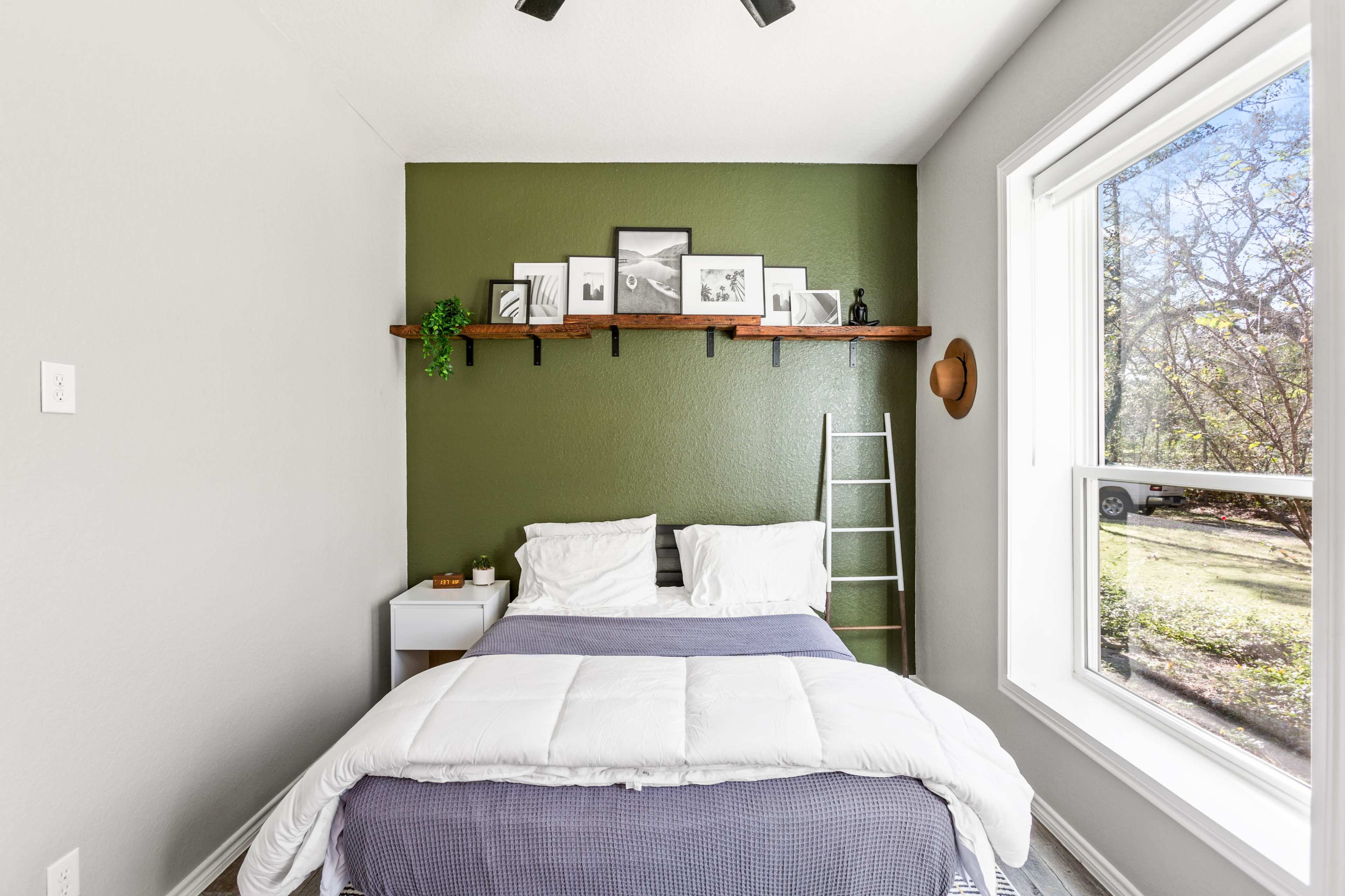 A small bedroom with a green accent wall, a double bed with white and gray bedding, a wooden shelf displaying framed pictures, a ladder, and a window overlooking a garden.