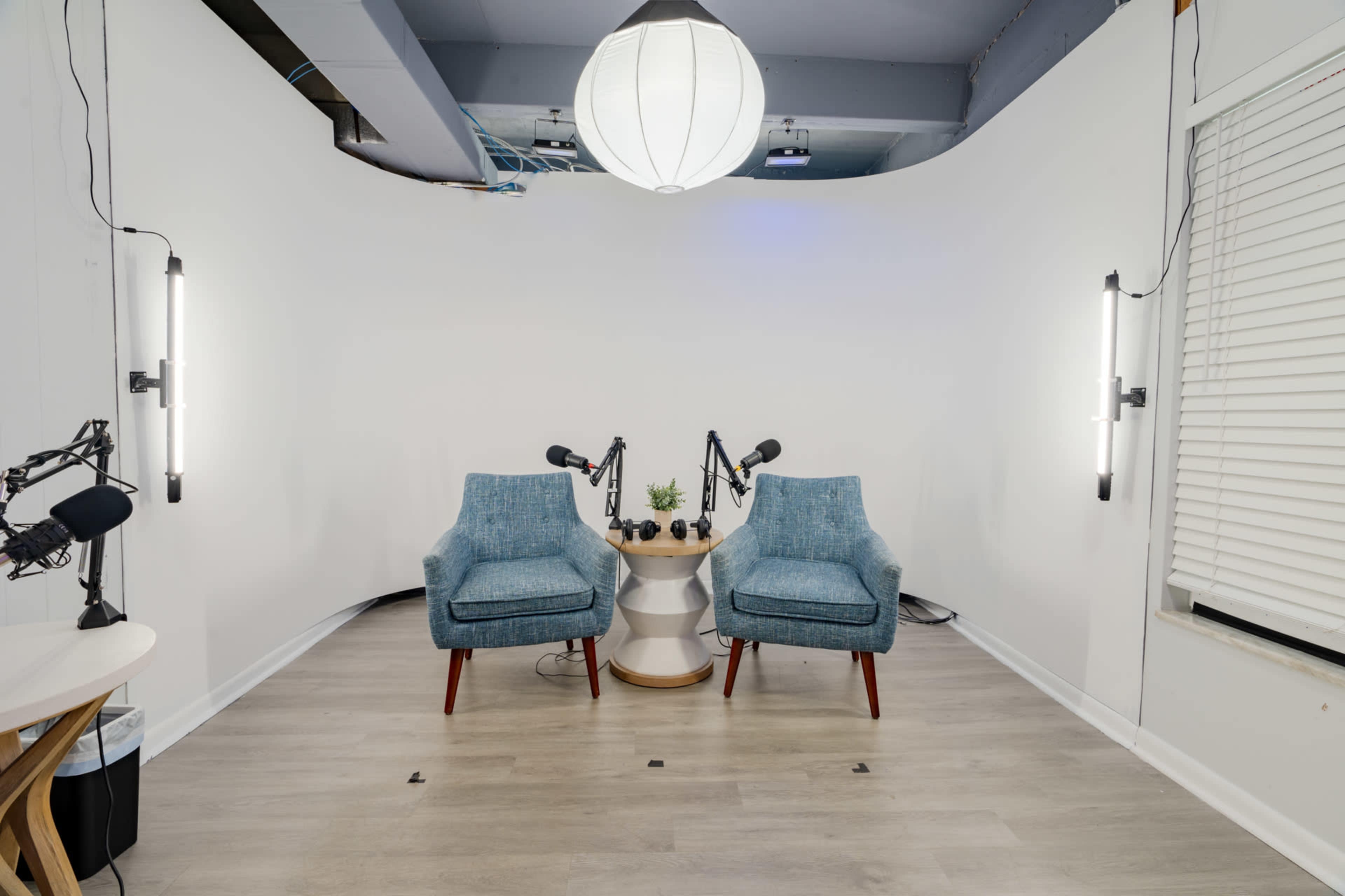 A minimalist recording studio with two blue chairs facing each other, a small round table between them, and microphones on stands attached to the walls.