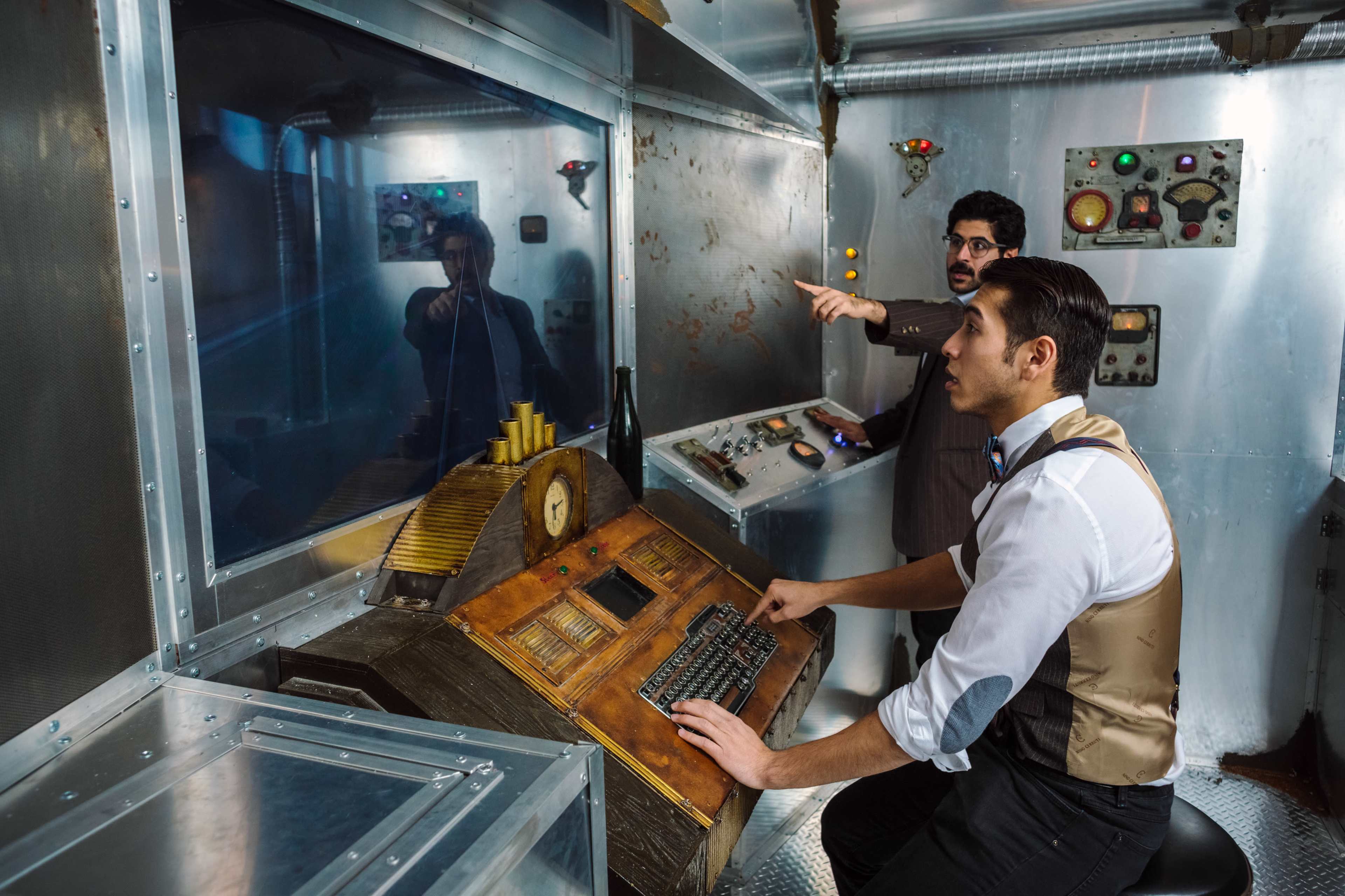 Two men are inside a metallic control room, one pointing at a screen while the other sits at a console with buttons and levers.