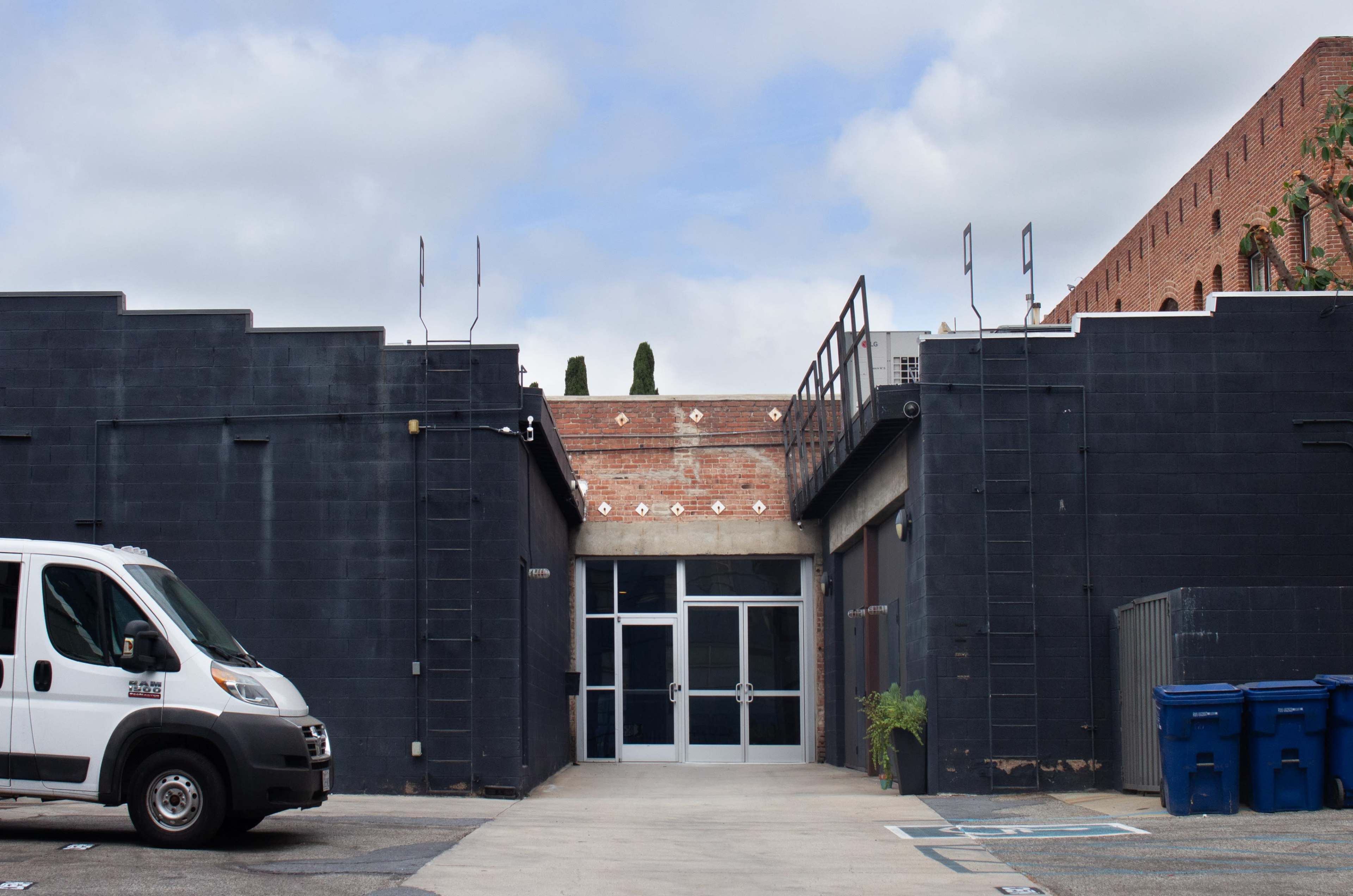 The image shows a narrow passageway between two black buildings, with a van parked on the left and glass doors at the end of the passage.