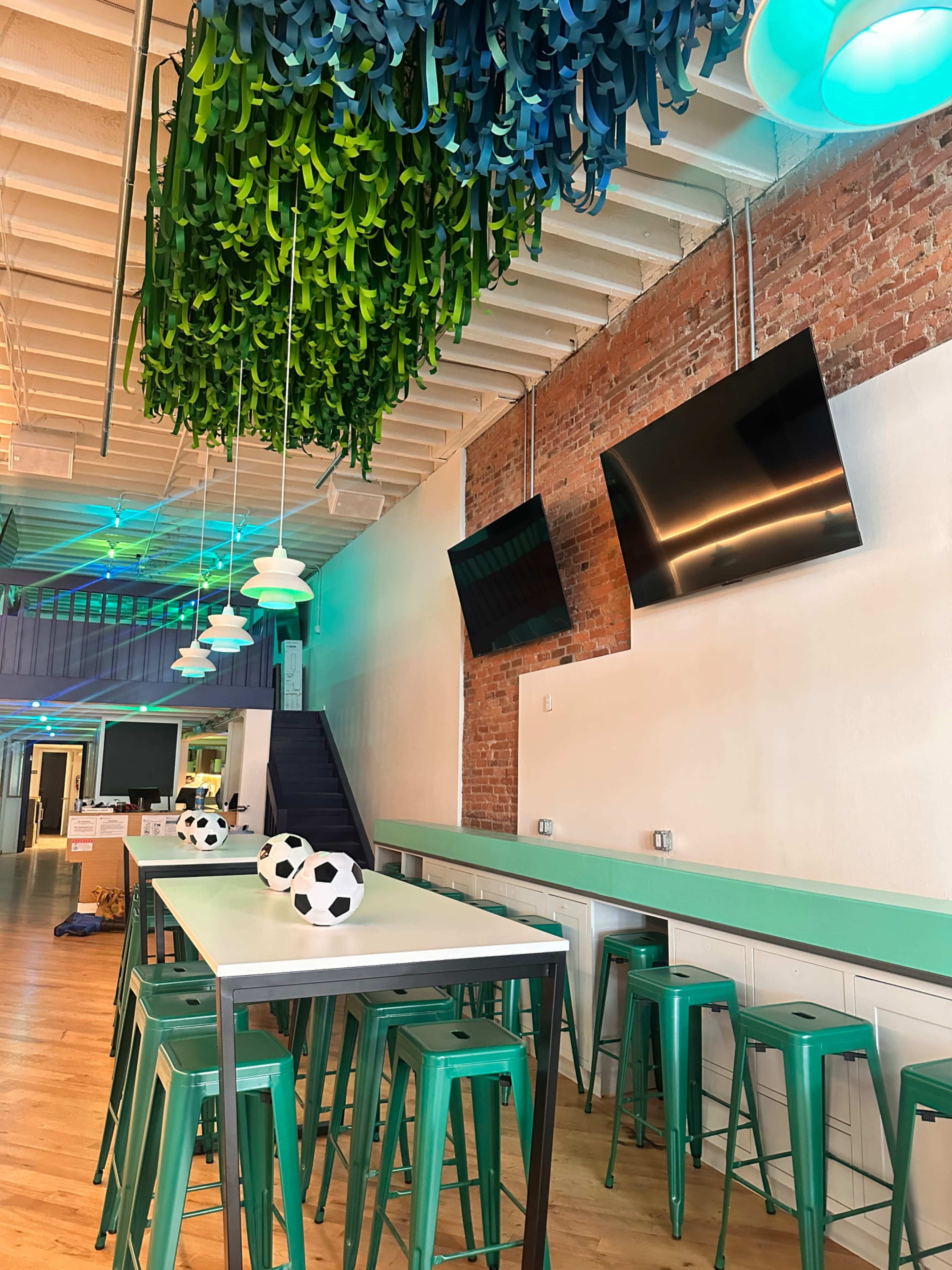The image shows a modern café interior with a long white table, green metal stools, and decorative hanging greenery overhead, along with two flat-screen TVs mounted on the wall.