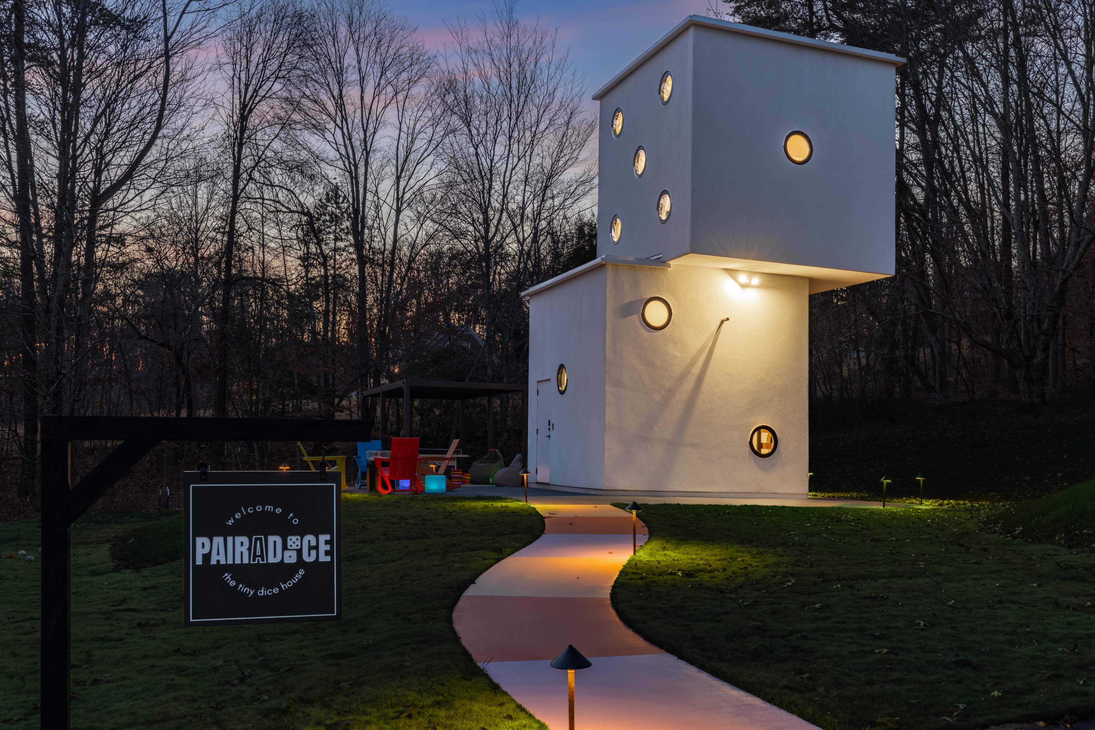 A unique, modern white structure with circular windows stands illuminated at dusk, accompanied by a path leading to a welcoming sign.