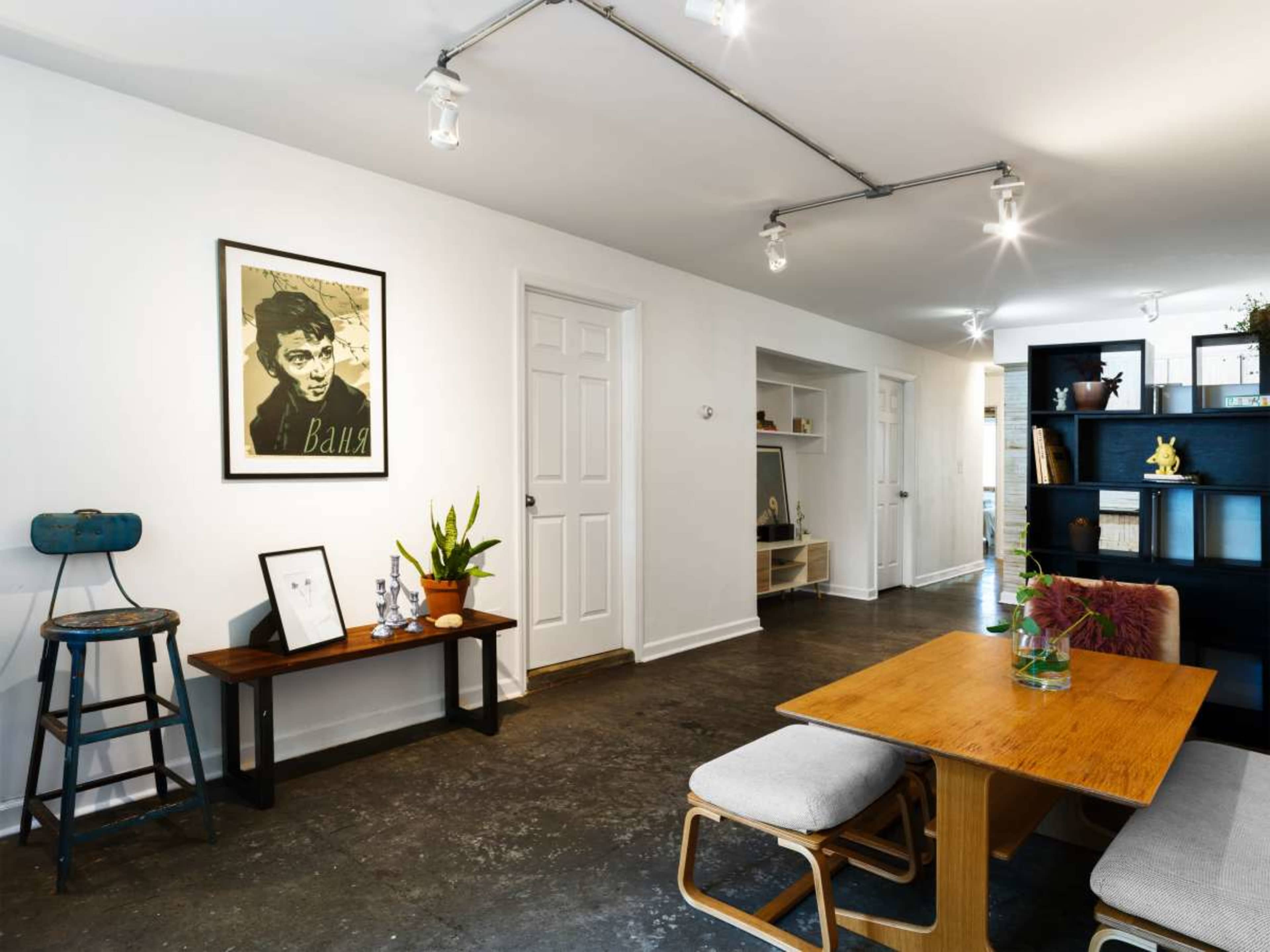 The image shows a minimalist interior space with a wooden table and chairs, a wall-mounted artwork, and a shelf holding various decorative items.