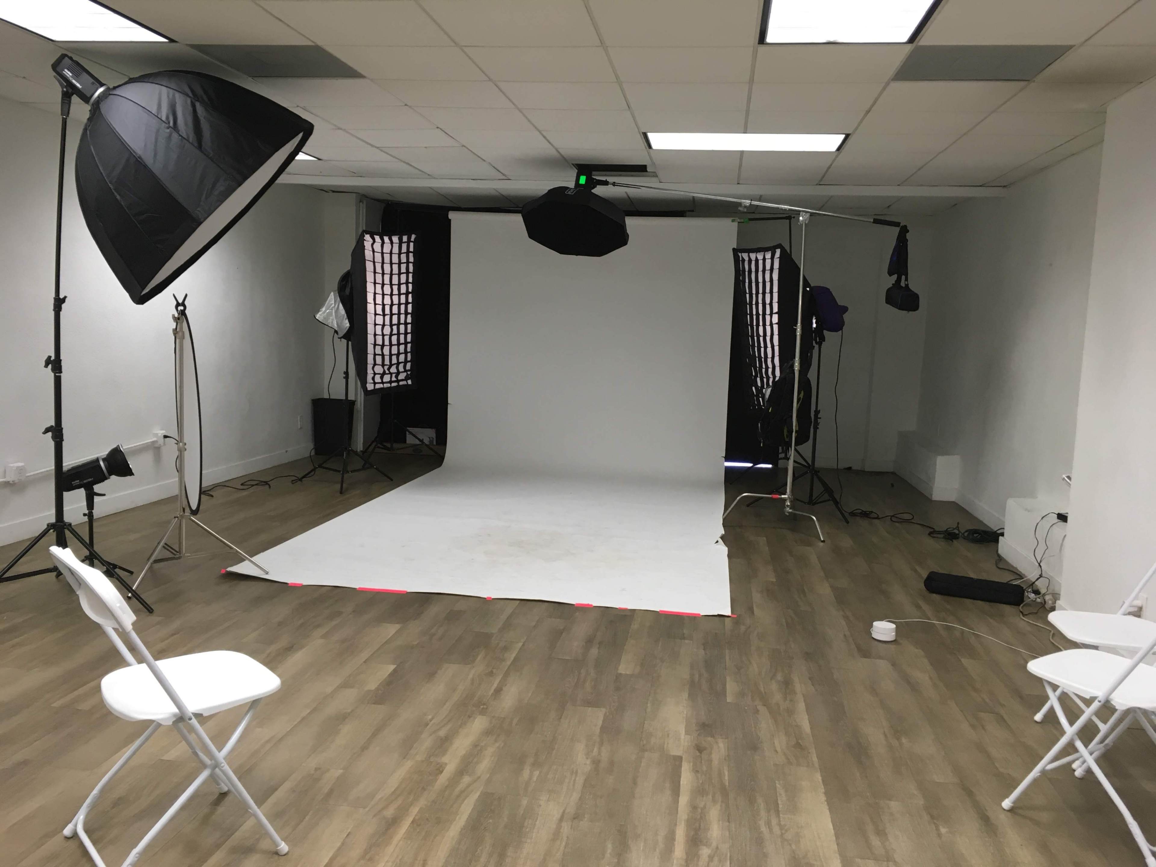 The image shows a photography studio with a white backdrop, lighting equipment, and several empty chairs arranged on a wooden floor.