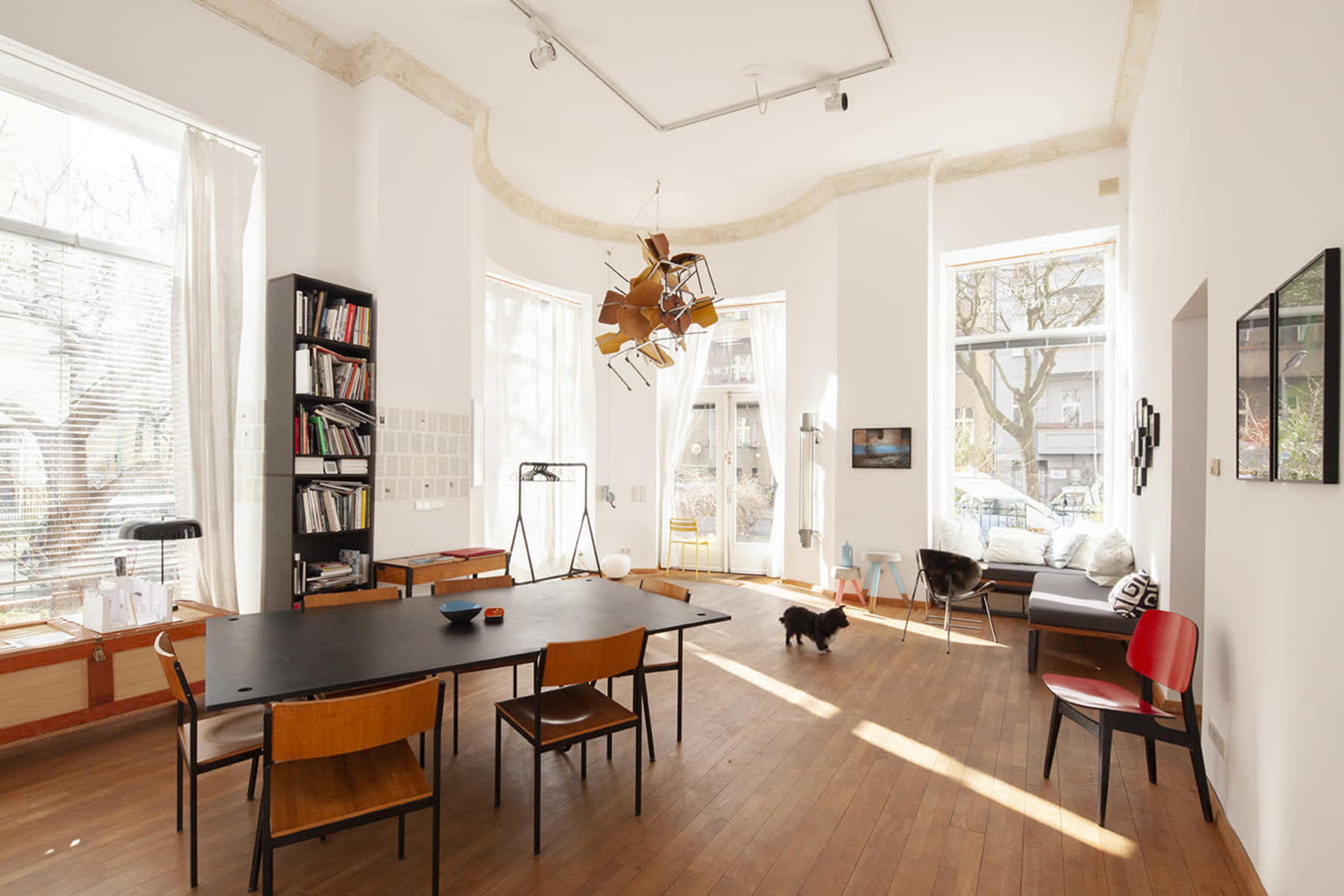 A spacious, well-lit room with large windows, a central dining table surrounded by chairs, and a bookshelf against one wall.