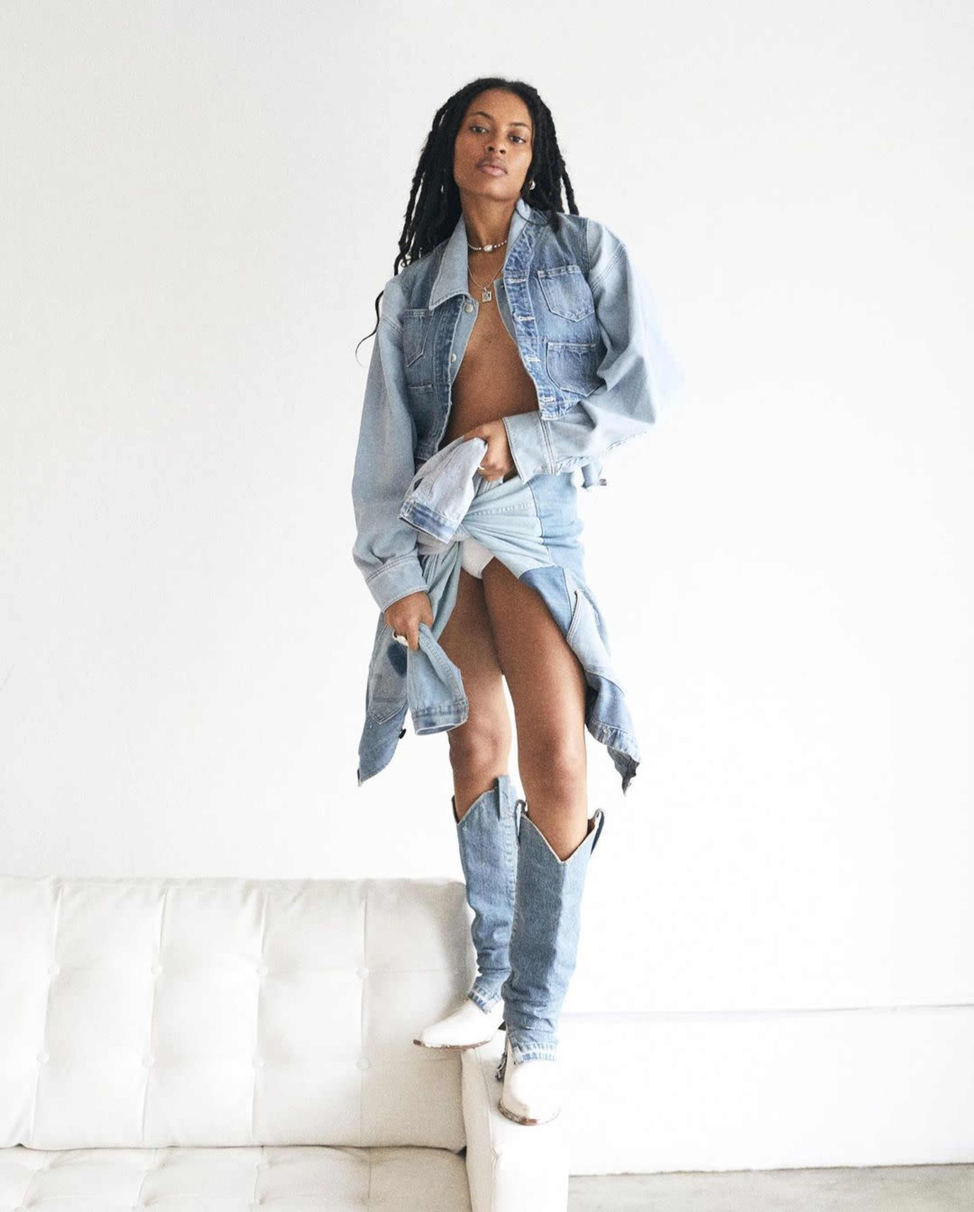 A woman with long, braided hair stands confidently on a white couch, wearing a denim jacket and a denim skirt with knee-high boots.