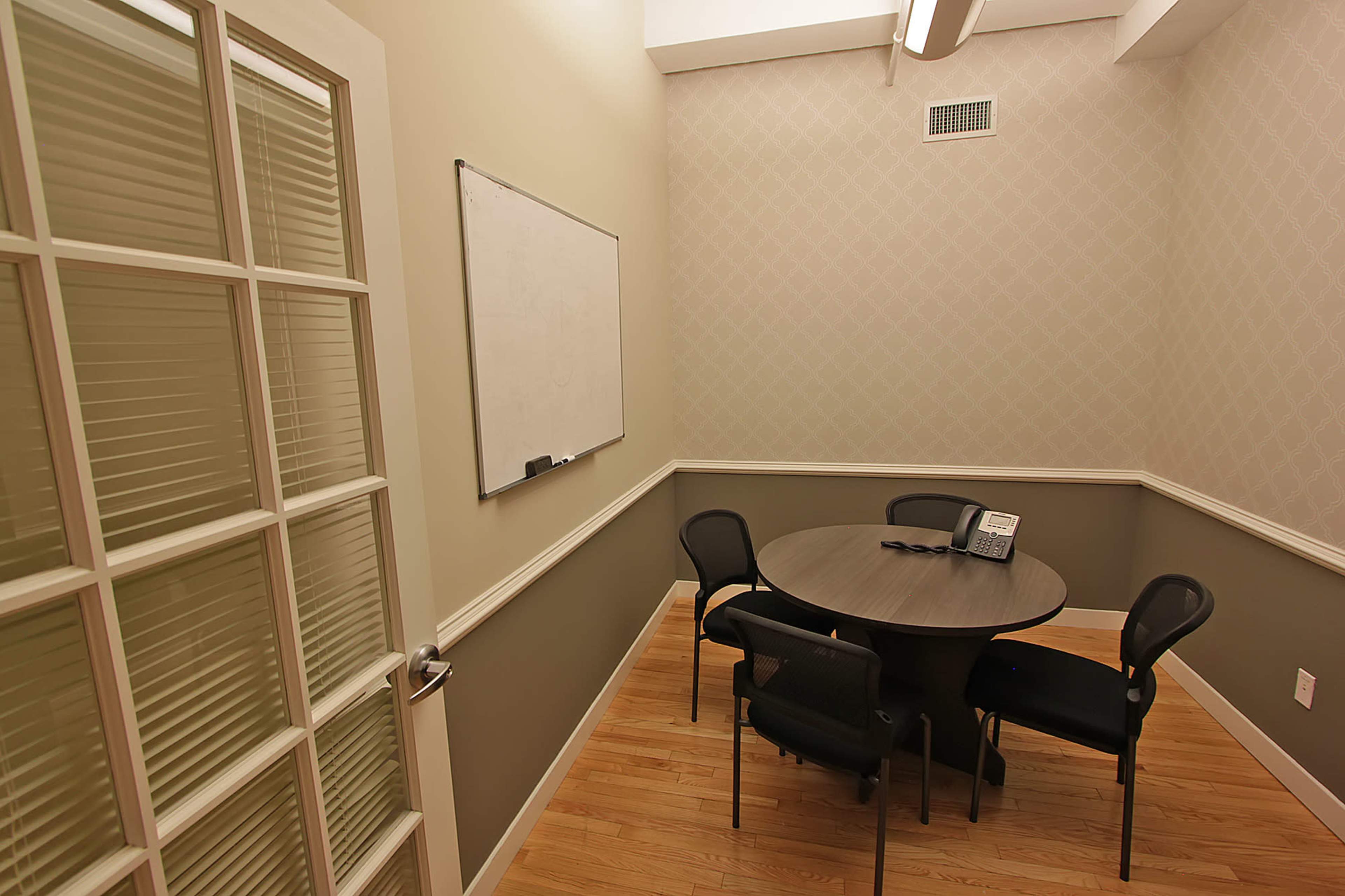 The image shows a small meeting room with a round table and four chairs, a whiteboard on the wall, and a telephone on the table.