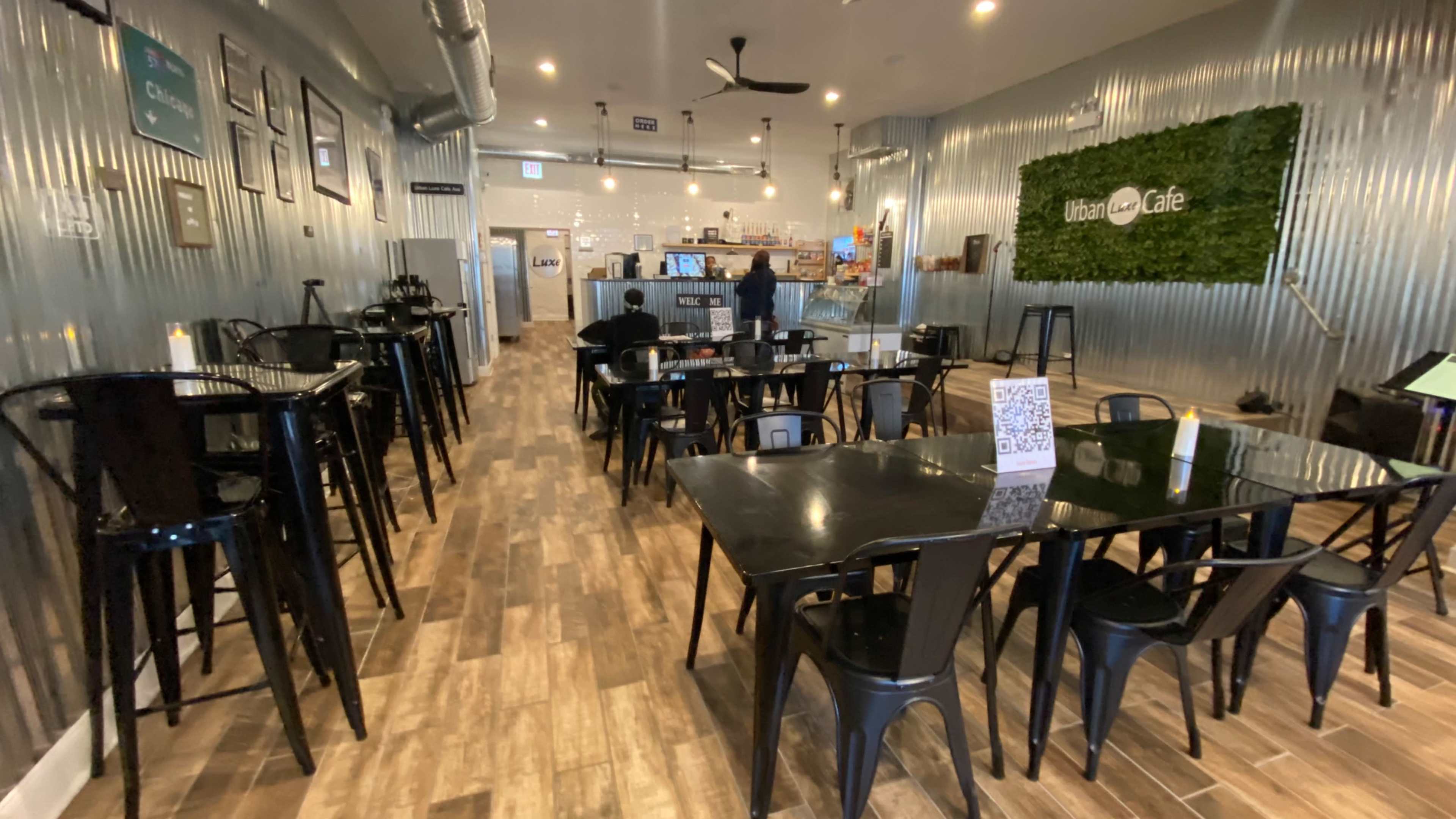 The interior of a café features black tables and chairs arranged neatly on a wooden floor, with a counter and kitchen area visible in the background.