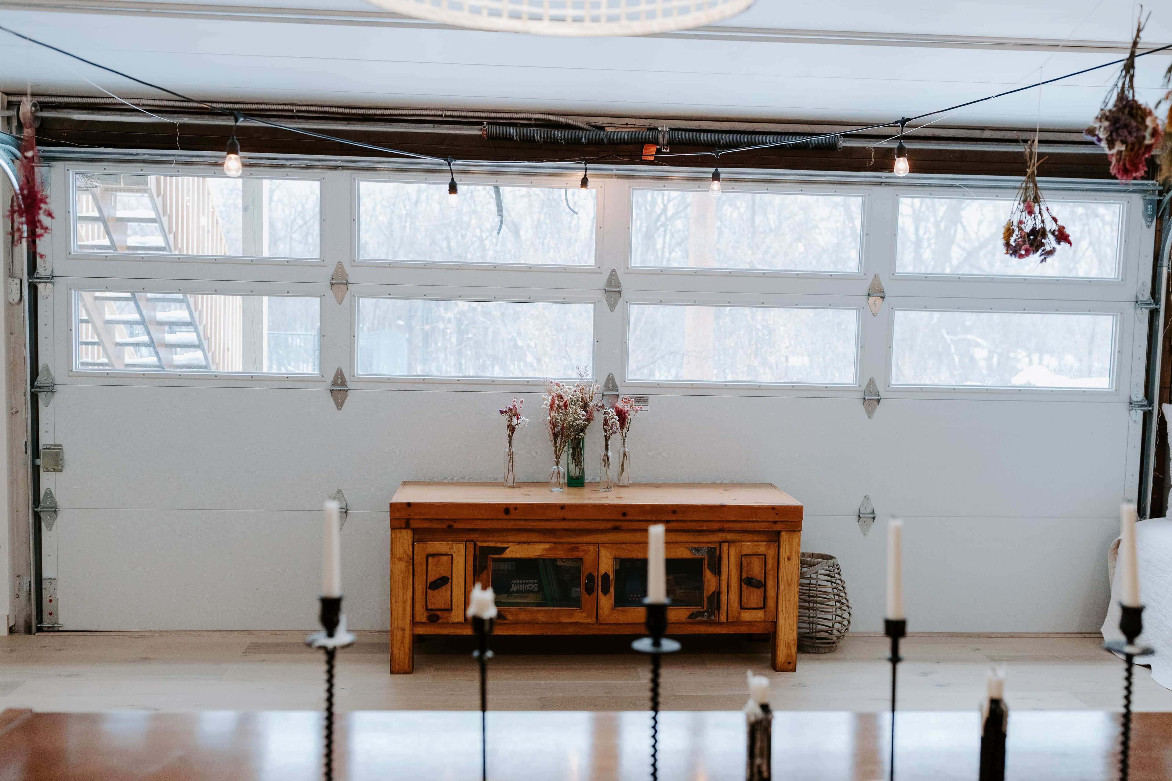 A spacious garage with large windows, a wooden console table displaying flowers, and light fixtures hanging from the ceiling.