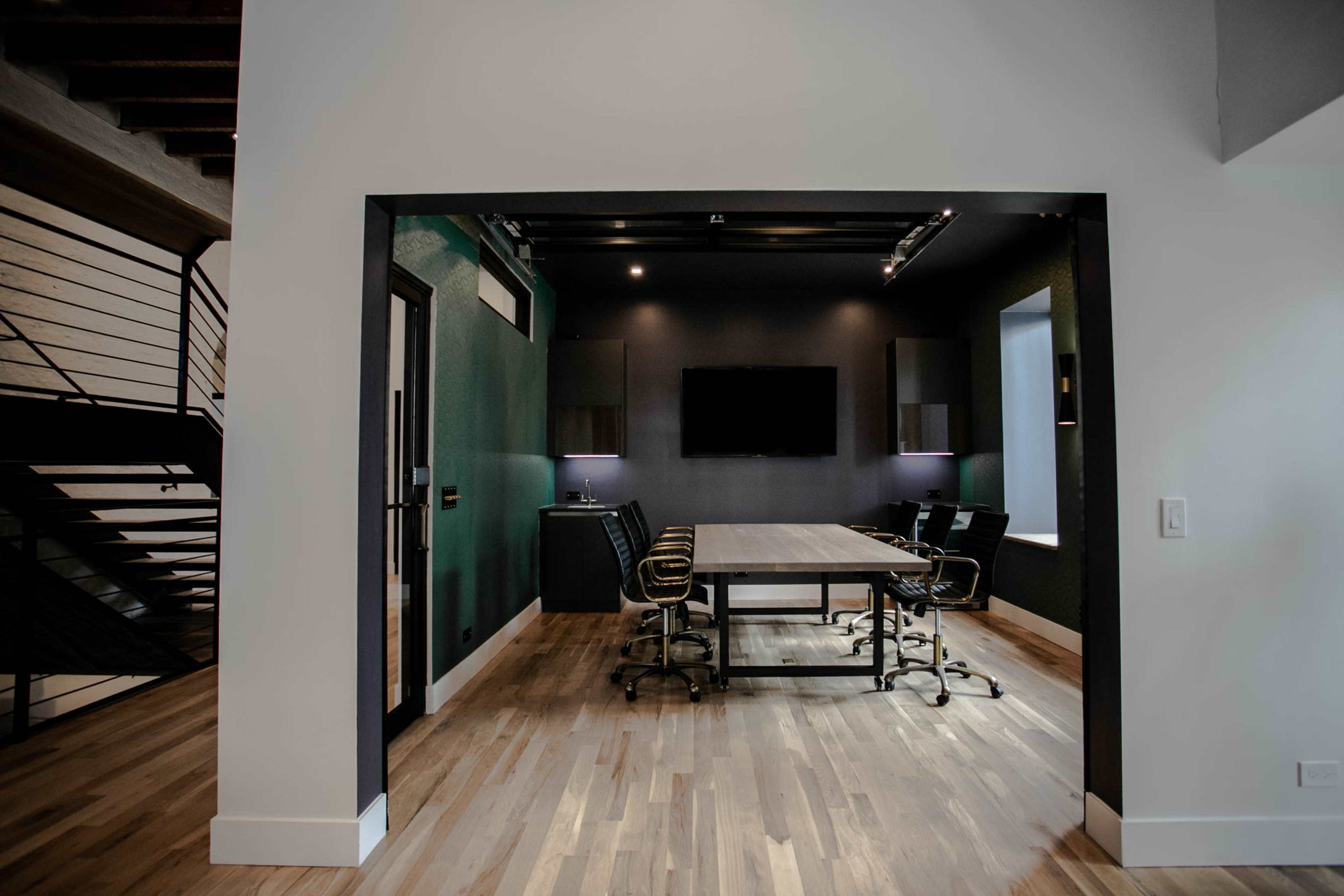 The image shows a modern conference room with a large table, black chairs, and a wall-mounted screen, viewed through an archway from an adjoining space.