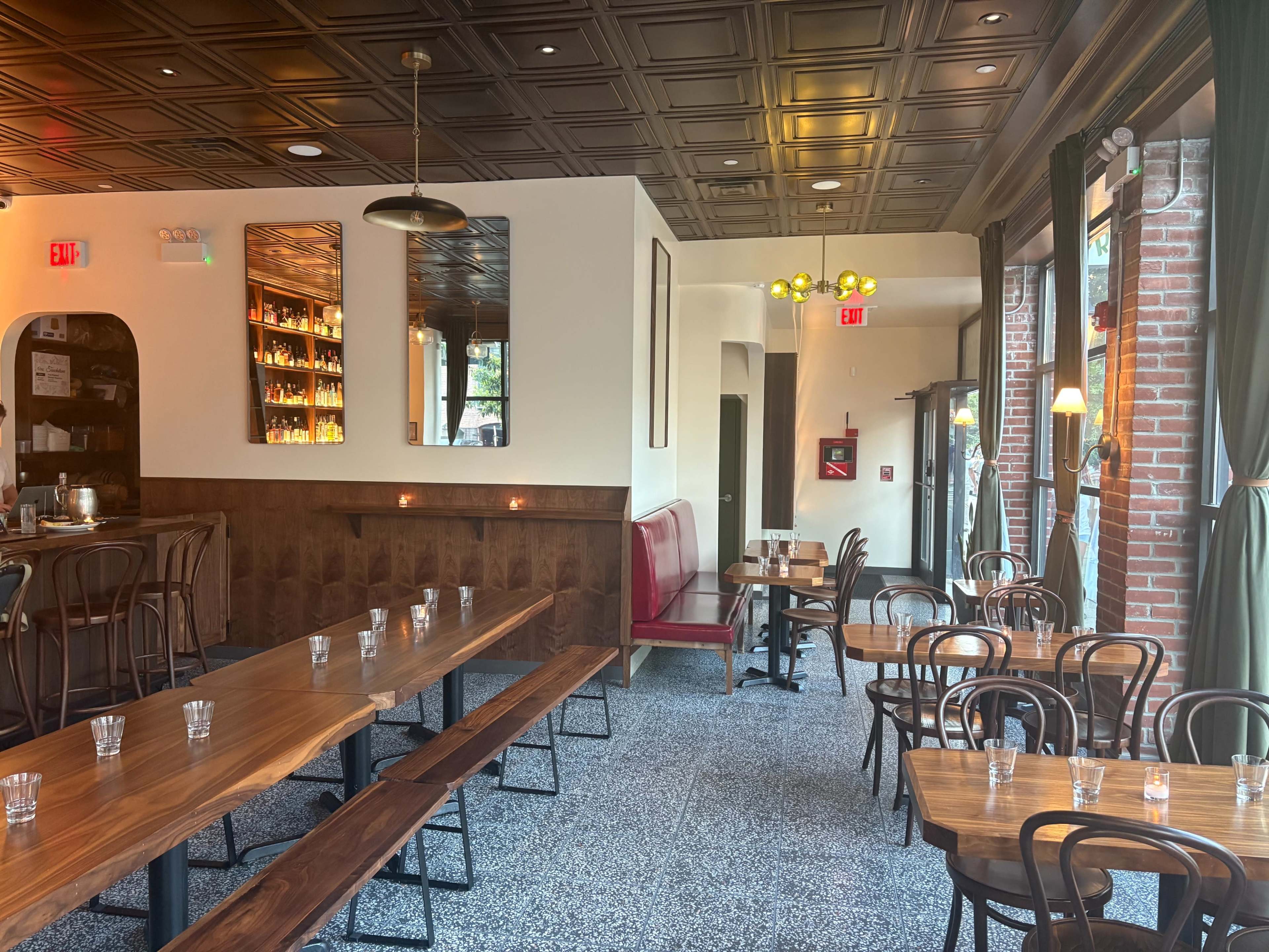 The image shows a modern restaurant interior featuring wooden tables, bentwood chairs, and wall-to-ceiling windows, with a bar area and decorative lighting.