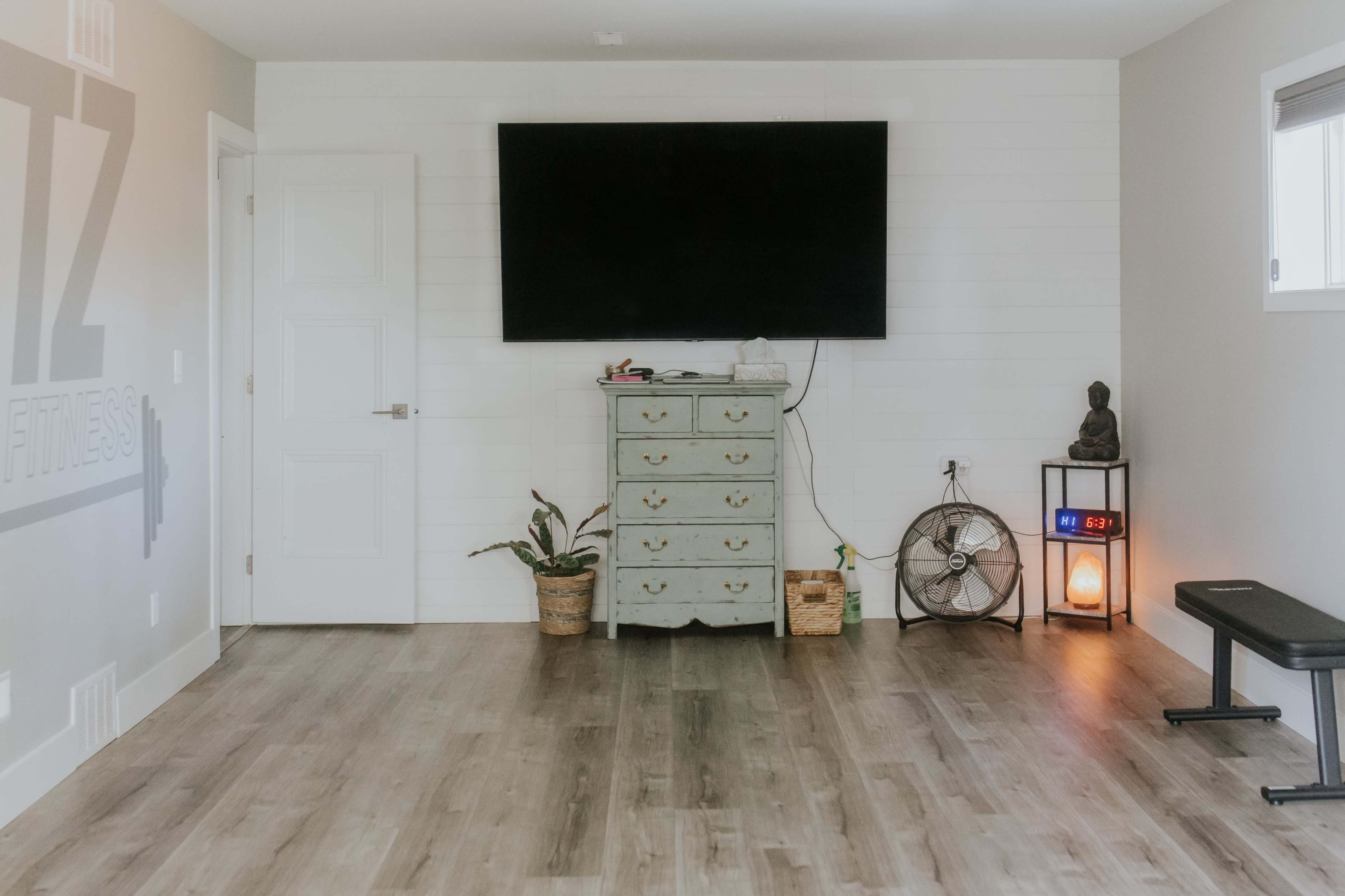 The image shows a minimalist fitness room featuring a large wall-mounted television, a green dresser, a fan, a small statue, and a bench against a light-colored wall.