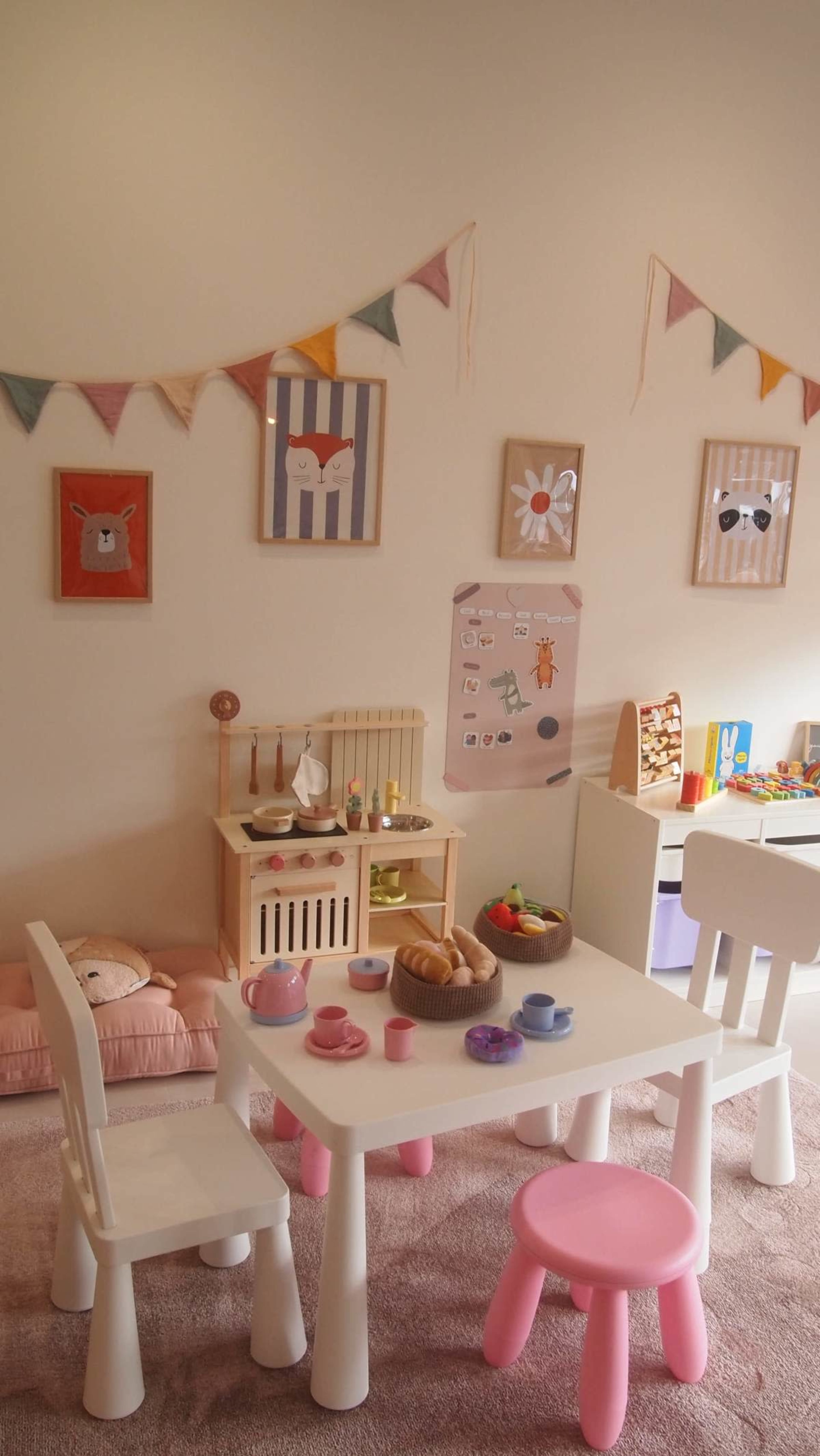 The image shows a children's playroom with a small table set with colorful toy dishes and food, surrounded by child-sized chairs and decorated walls featuring playful artwork and bunting.