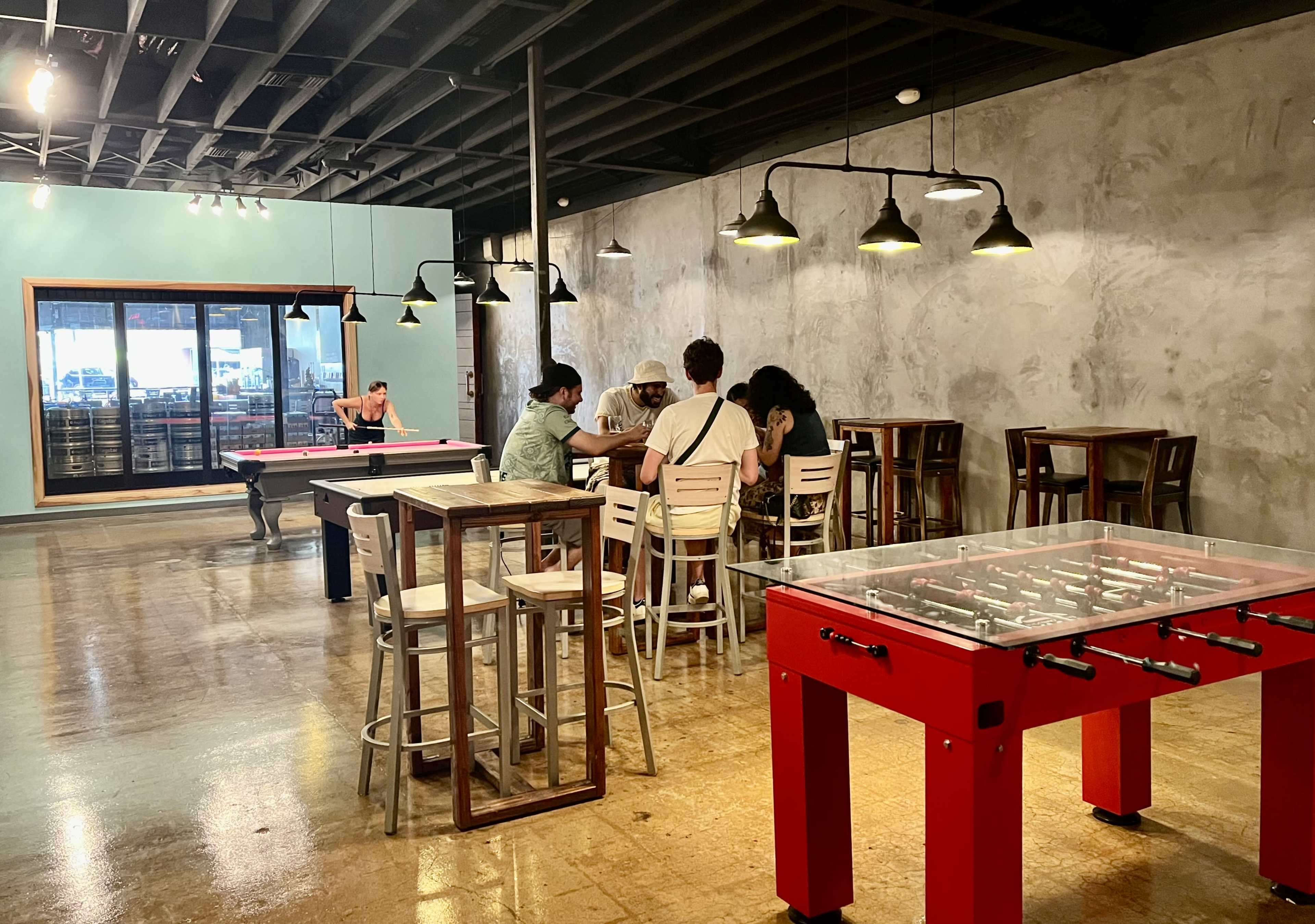 The image shows a casual interior space with pool tables, a foosball table, and a group of people seated at a table under pendant lights.