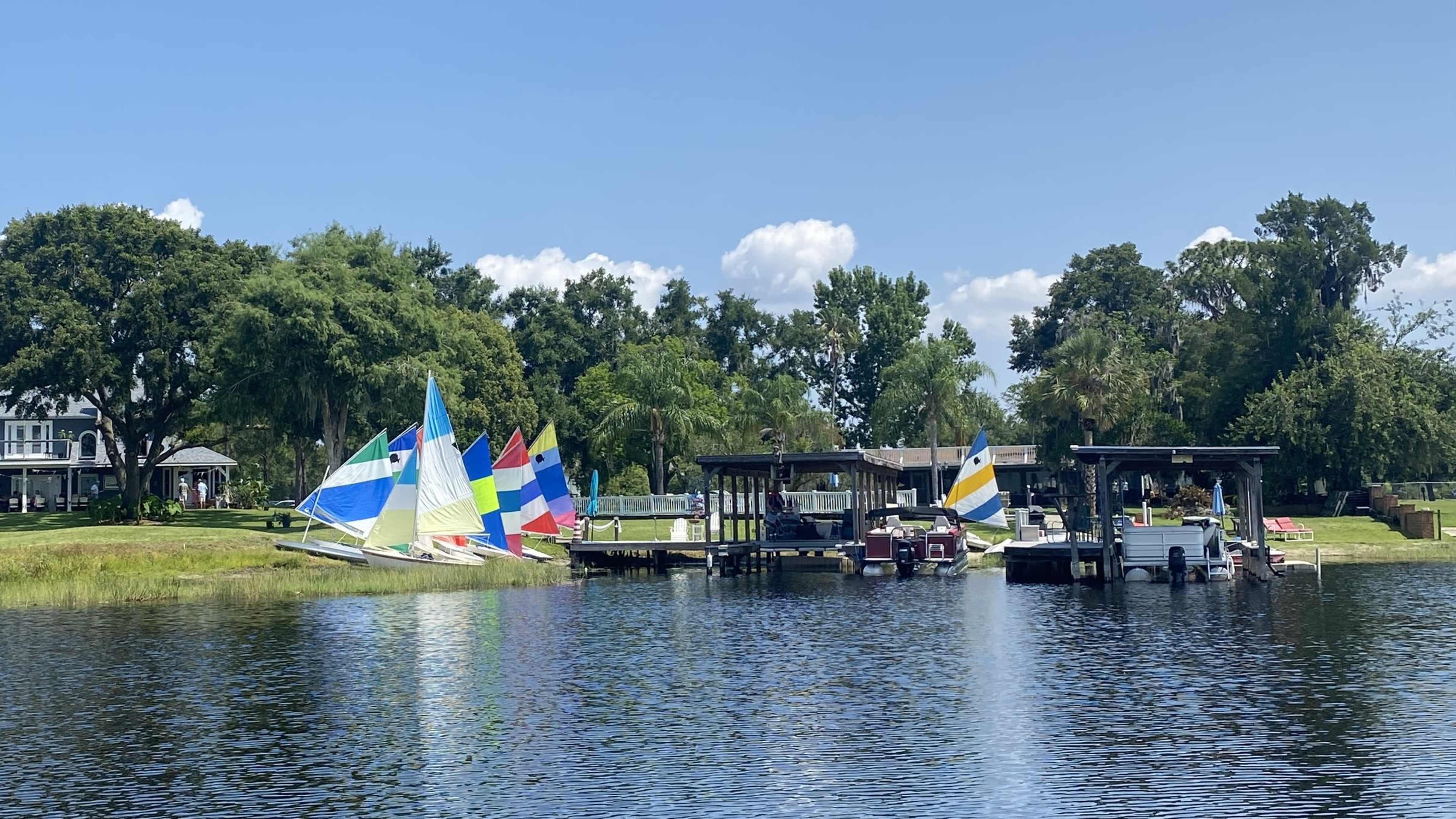 Sailboats with colorful sails are moored near docks on a calm body of water surrounded by greenery.