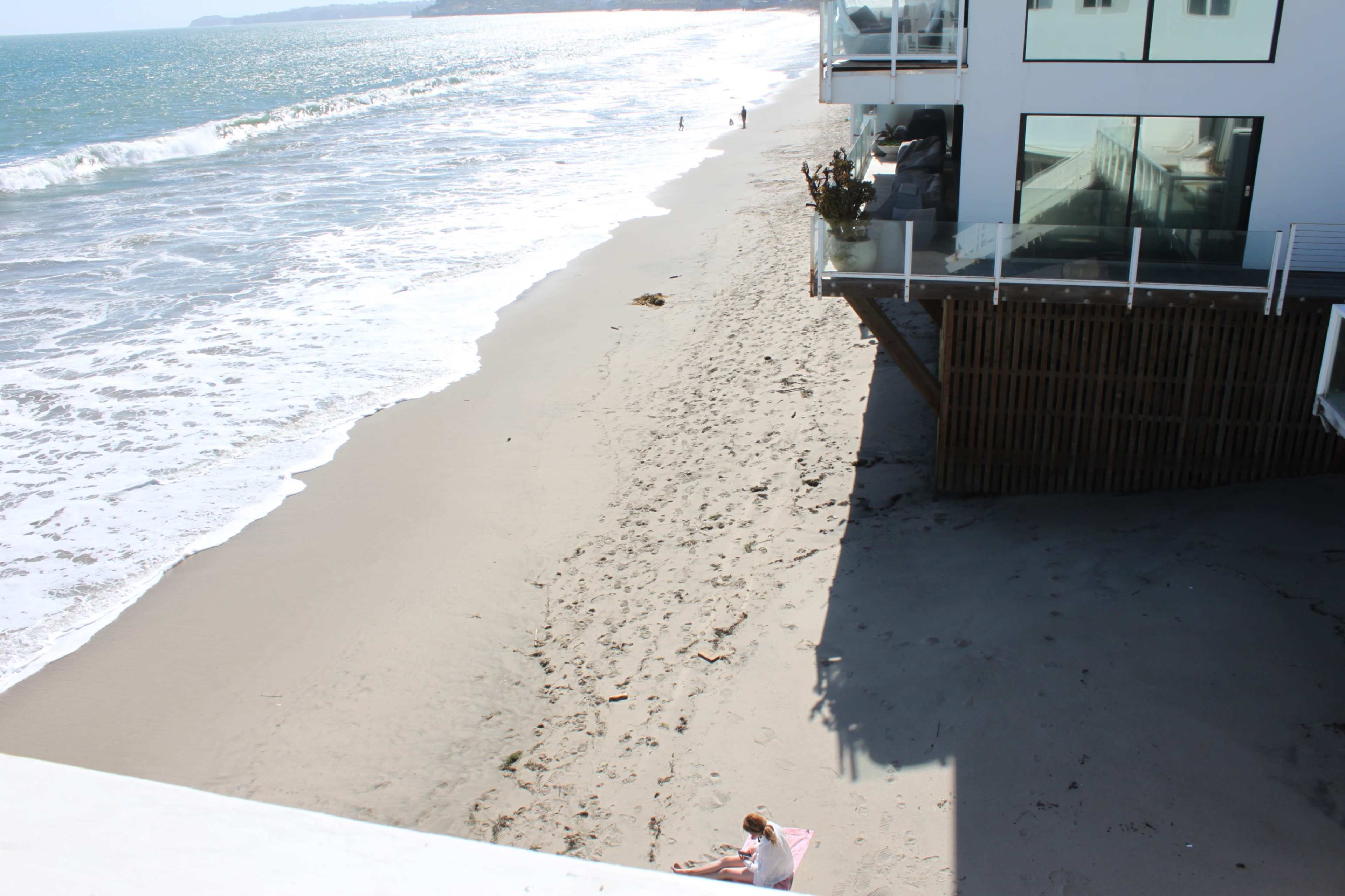 The image shows a sandy beach beside a shoreline with ocean waves, while a person sits on the sand and another walks along the water's edge near a modern building.