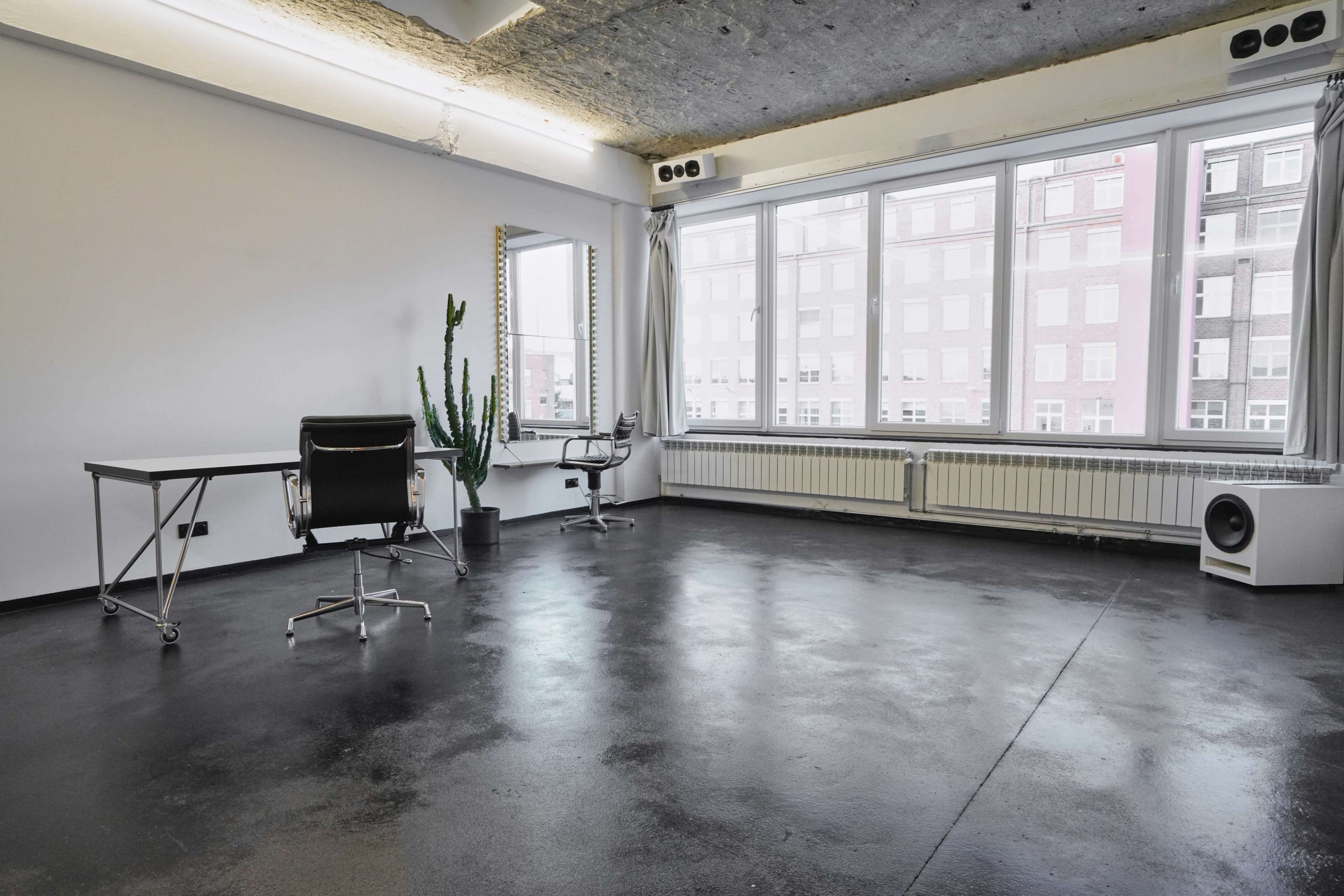 The image shows a minimalist office space with a desk, a chair, a potted cactus, and large windows that provide natural light.
