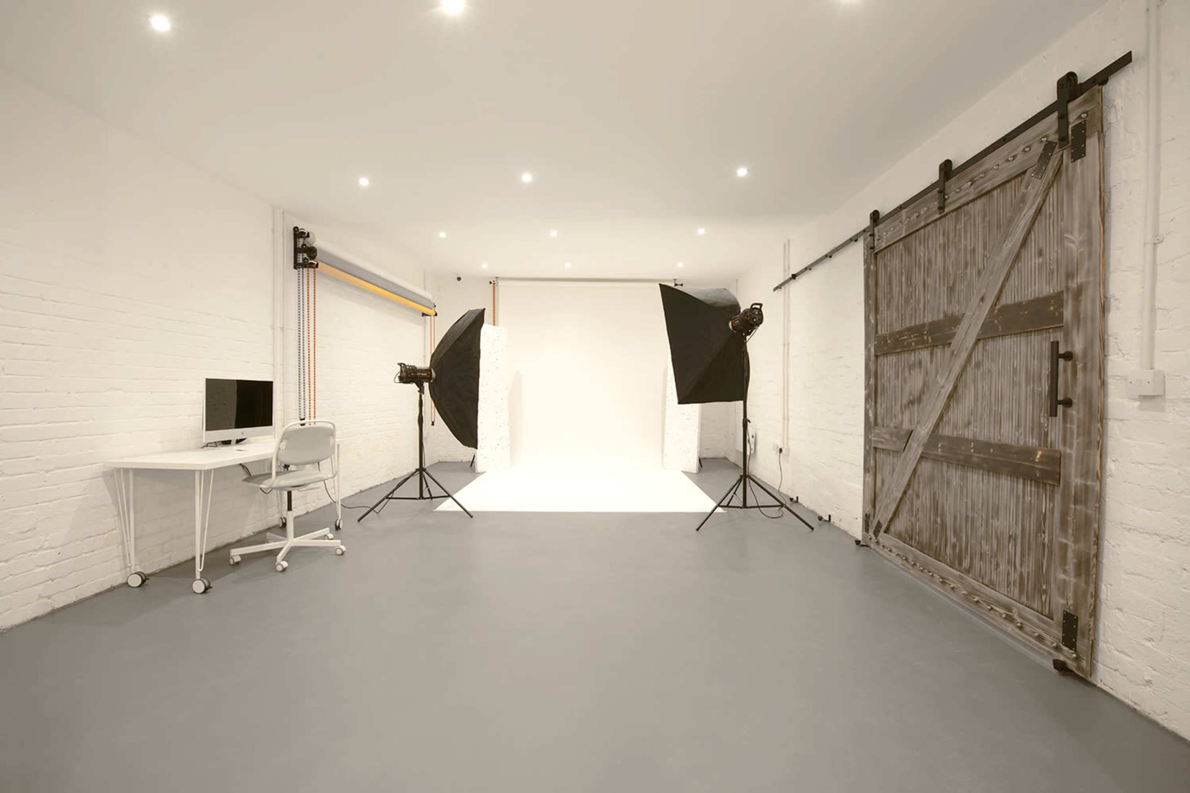 A spacious photography studio featuring a white backdrop, studio lighting, a desk with a computer, and a sliding barn door against a white brick wall.