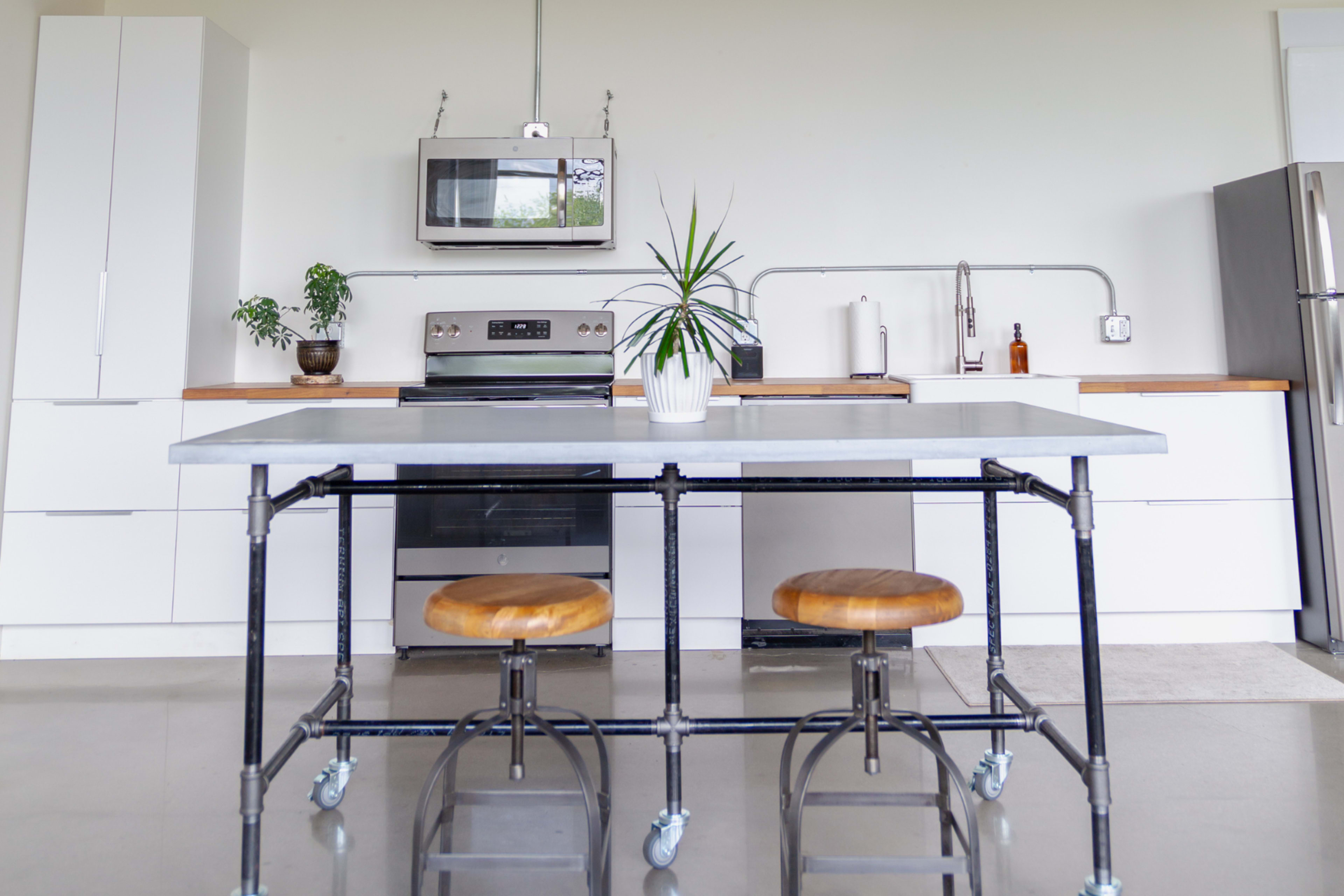 A modern kitchen features a metal-framed table with wooden stools, stainless steel appliances, and minimalist cabinetry.
