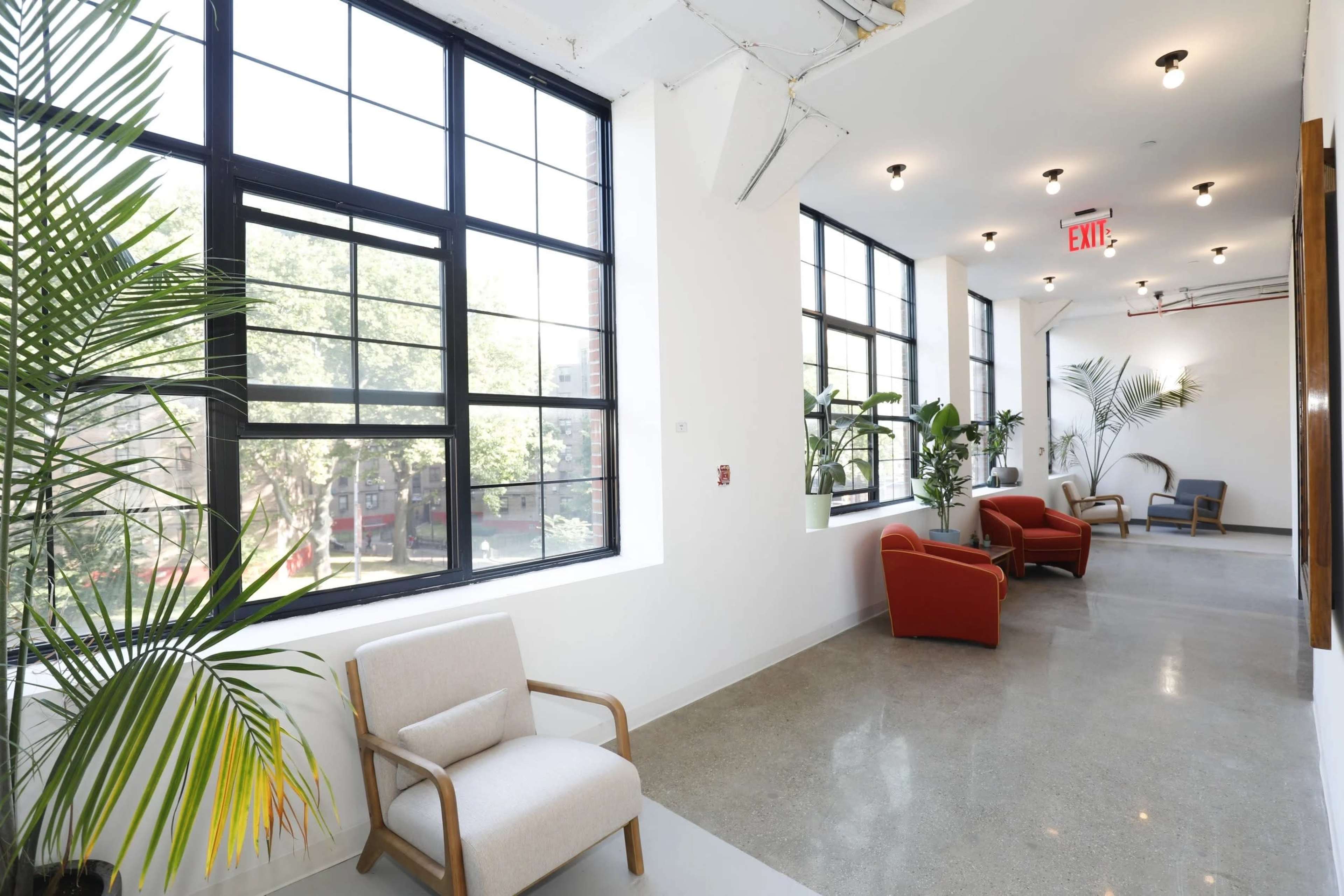 A hallway features large windows, potted plants, and a mix of armchairs along the walls.