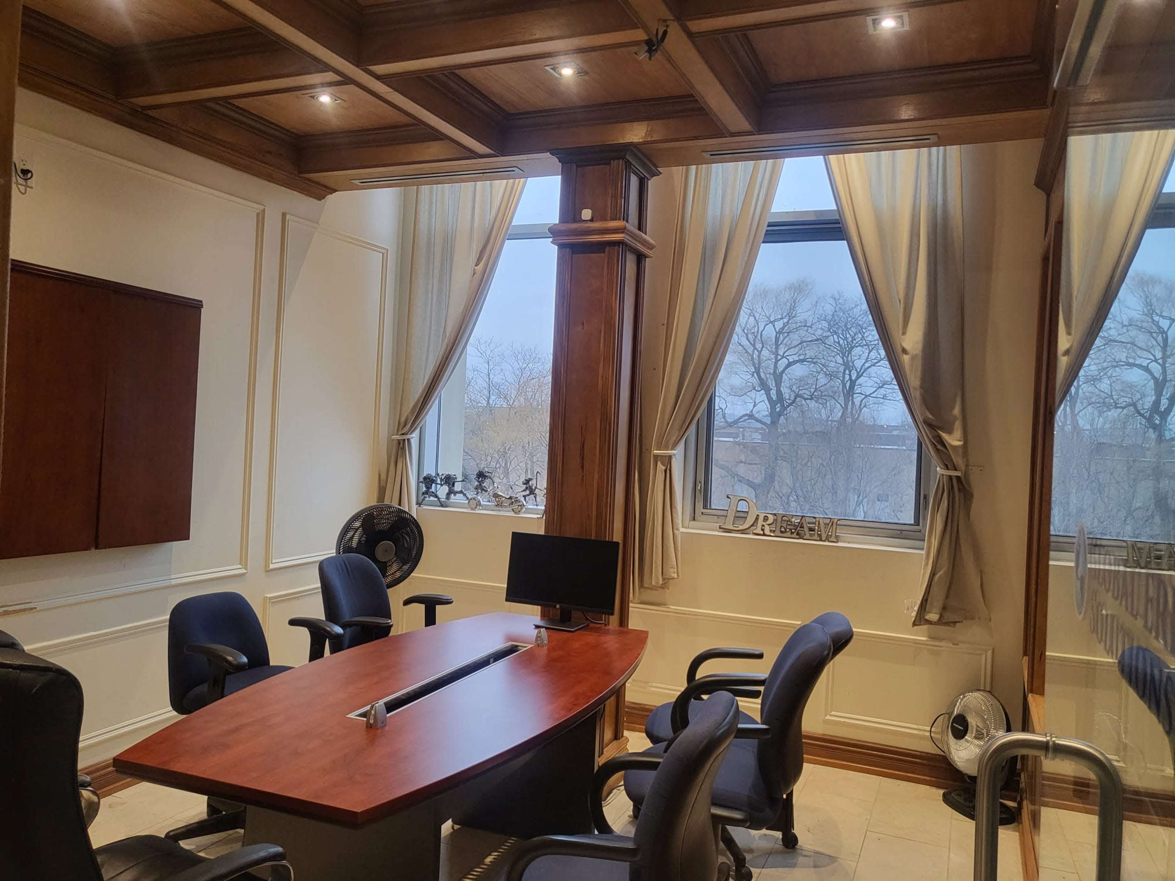 A conference room features a wooden table, several black chairs, and large windows with sheer curtains, revealing a view of bare trees outside.
