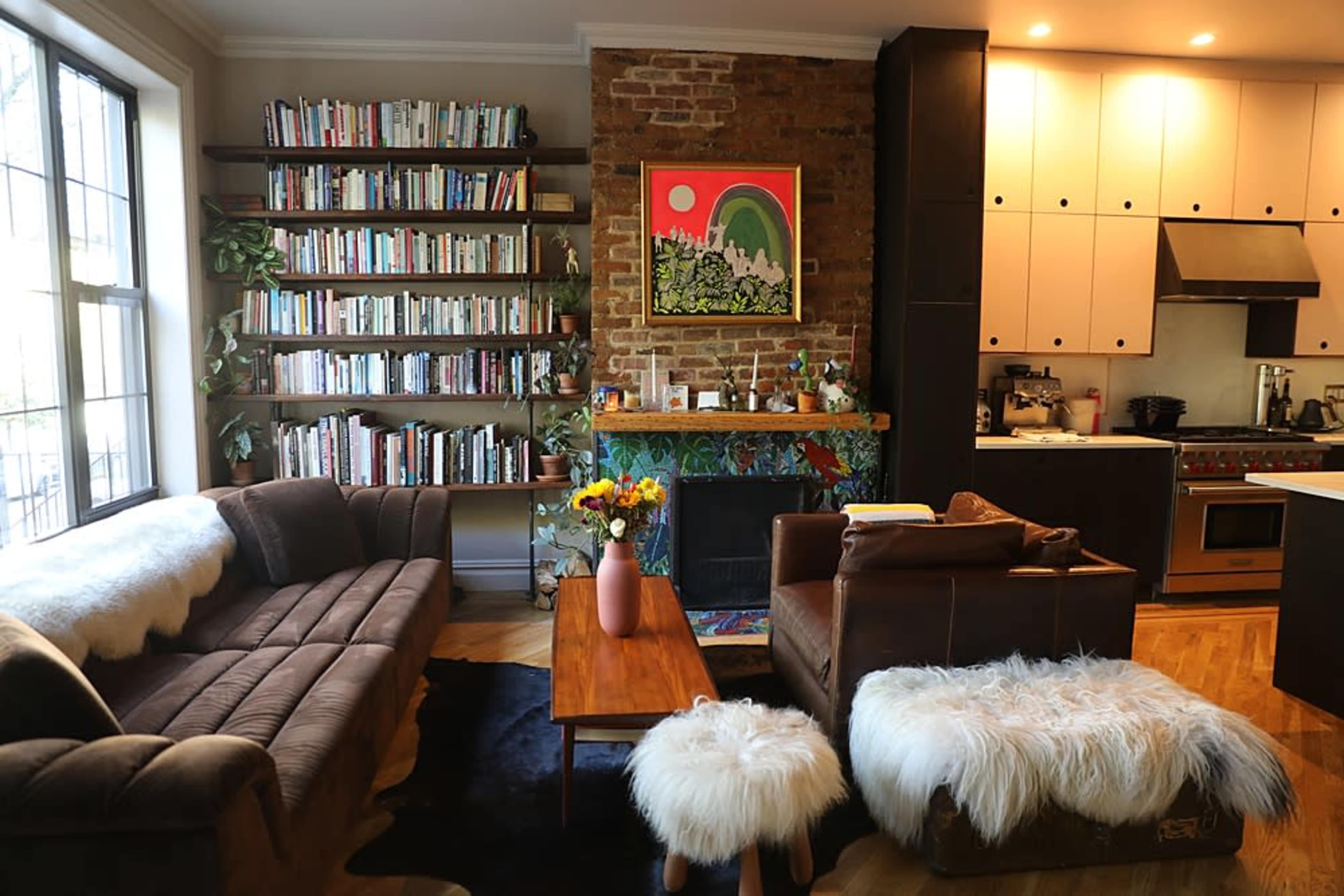 The image shows a modern living room featuring a brown sectional sofa, a wooden coffee table, and shelves filled with books, alongside a brick wall and a kitchen area in the background.