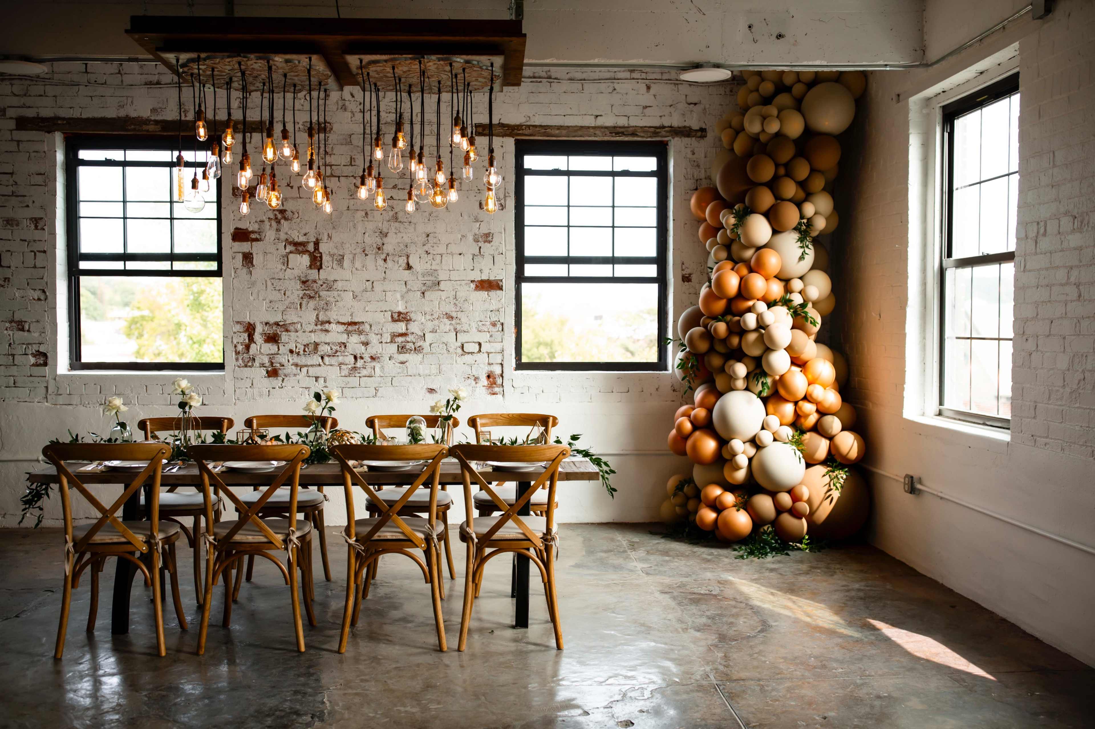 A large dining table with wooden chairs is set in a brightly lit room featuring exposed brick walls and a decorative arrangement of balloons in warm tones against one wall.