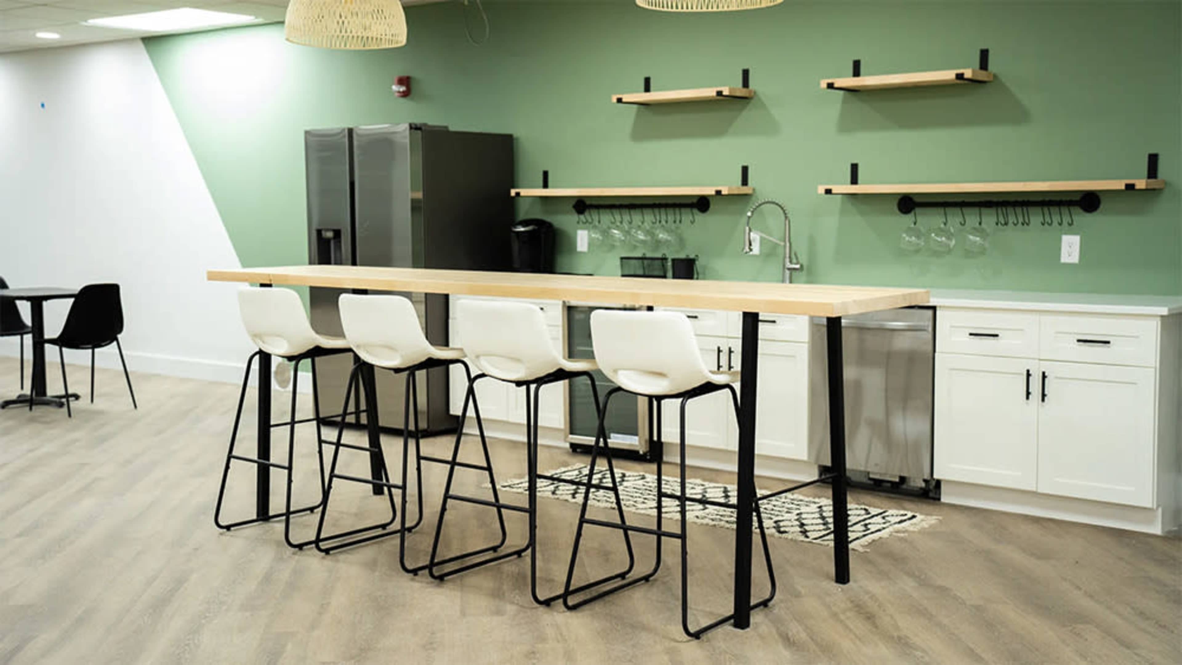 A kitchen area features a long wooden counter with four high stools, a green wall, and shelves above for storage.
