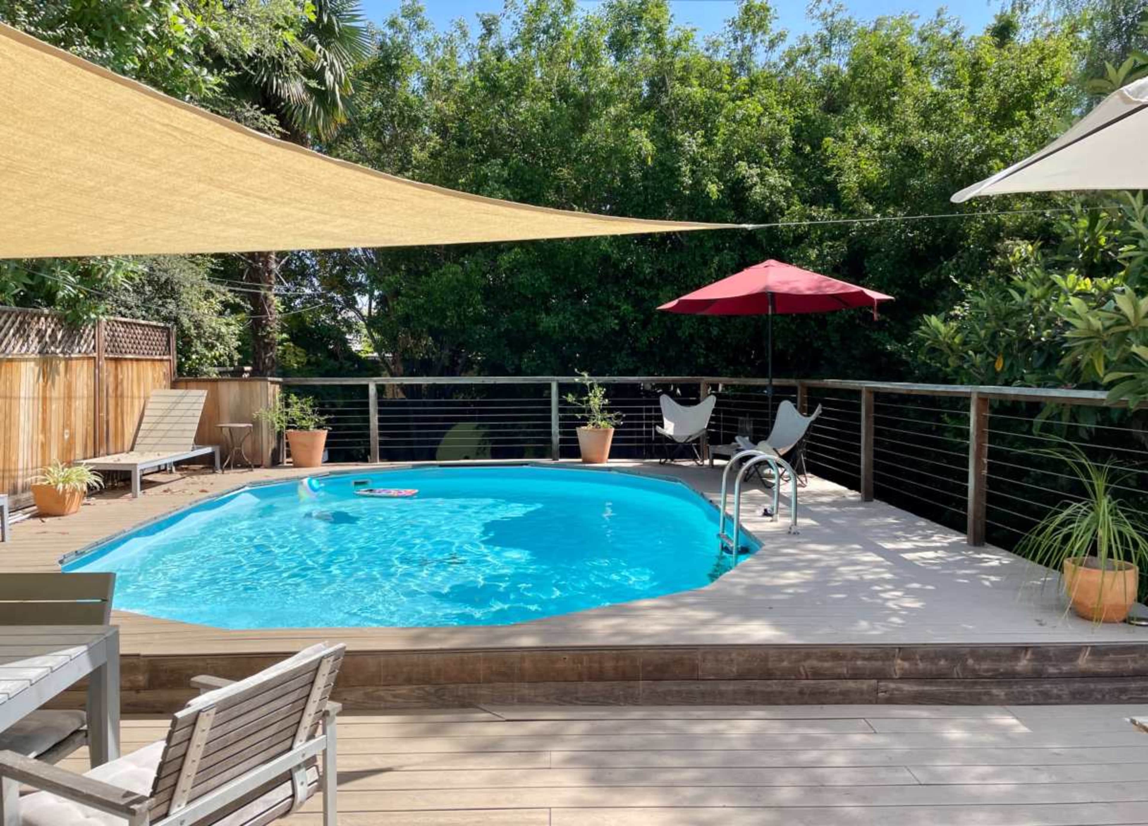 The image shows a backyard with a blue pool surrounded by a wooden deck, lounge chairs, potted plants, and shade sails overhead.