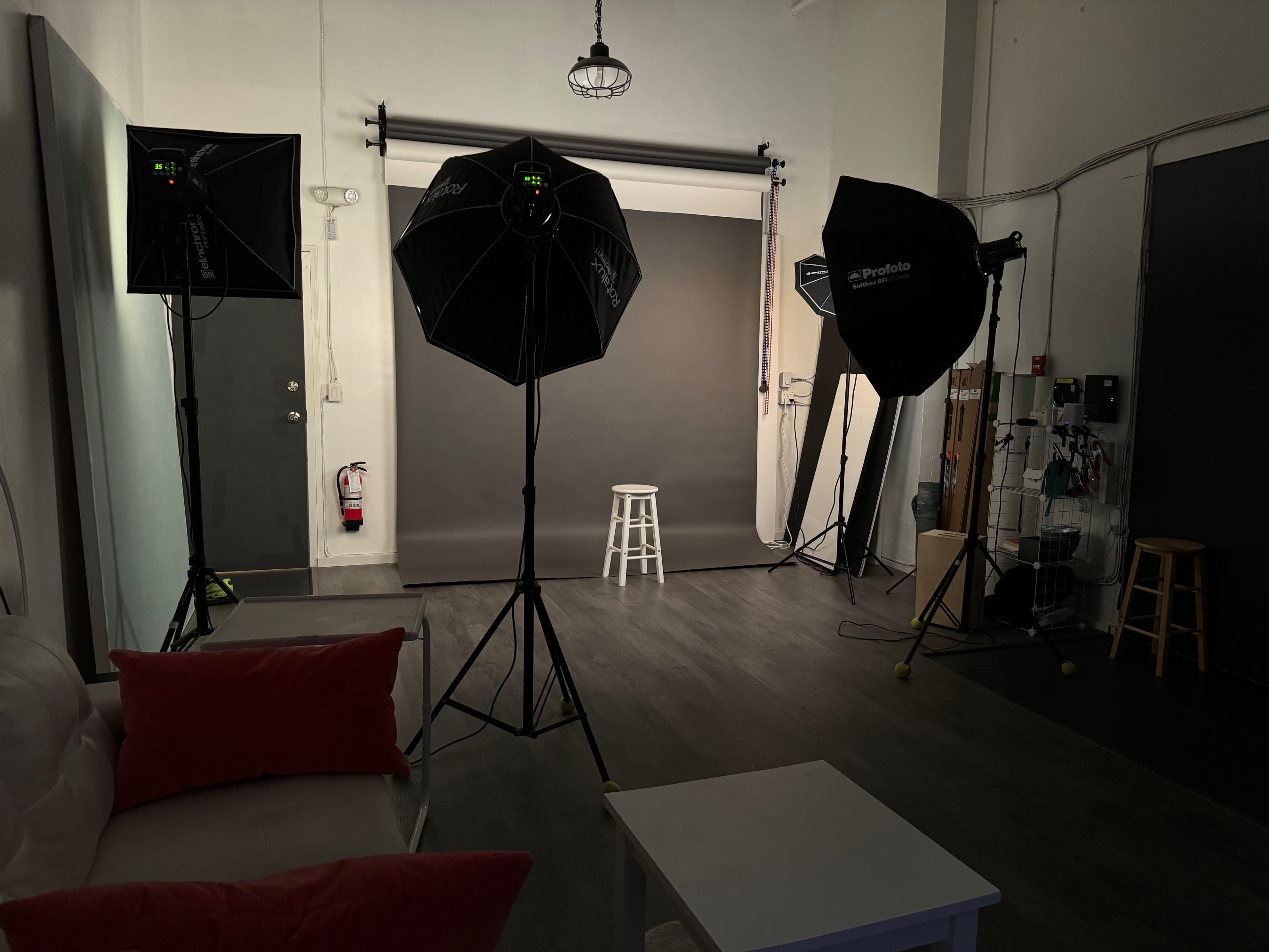 A photography studio features a gray backdrop, a stool in the center, and multiple softbox lights arranged around it.