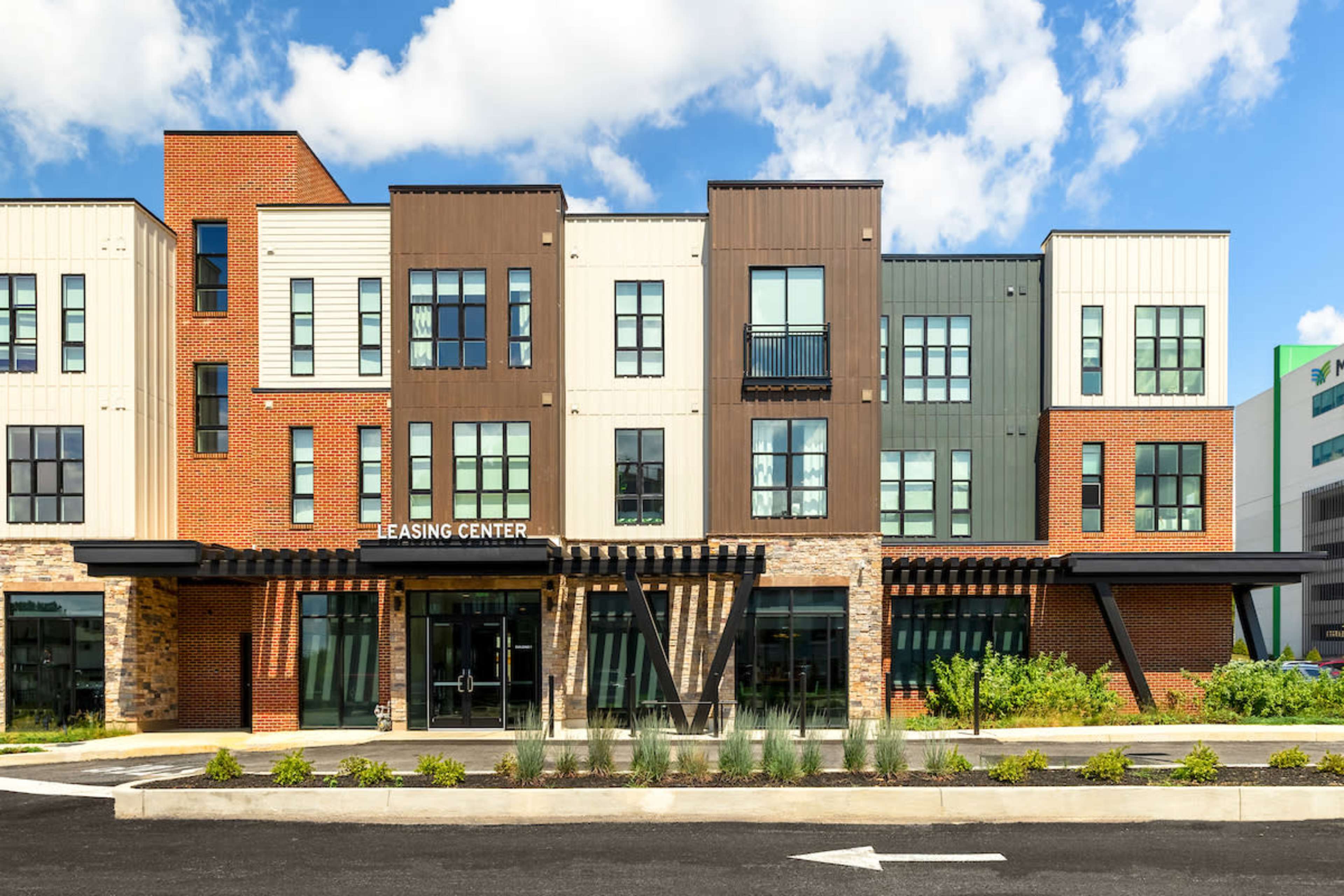 The image shows a modern leasing center building with a mix of brick and metal siding, featuring multiple windows and a covered entrance.