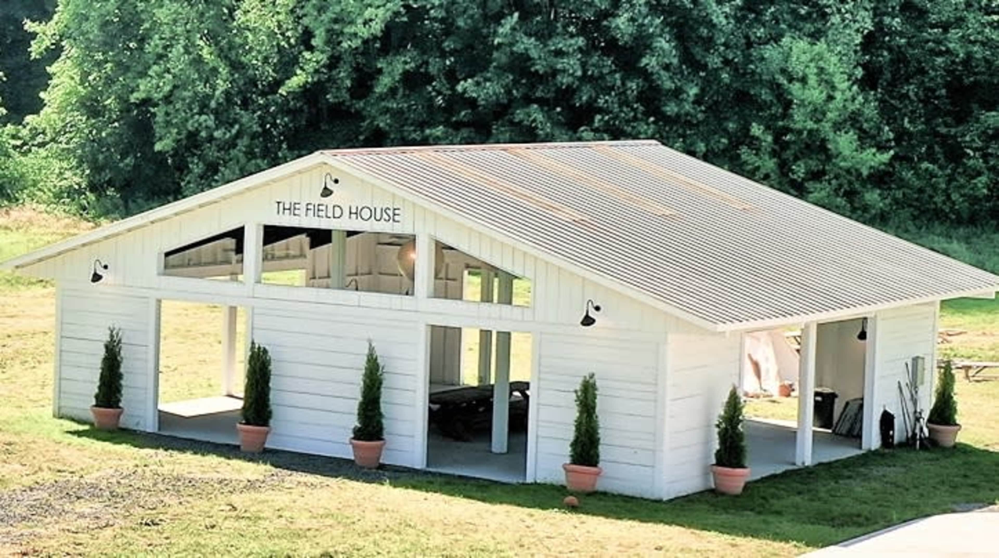 A white wooden building with a metal roof, labeled "The Field House," surrounded by greenery and decorative potted plants.