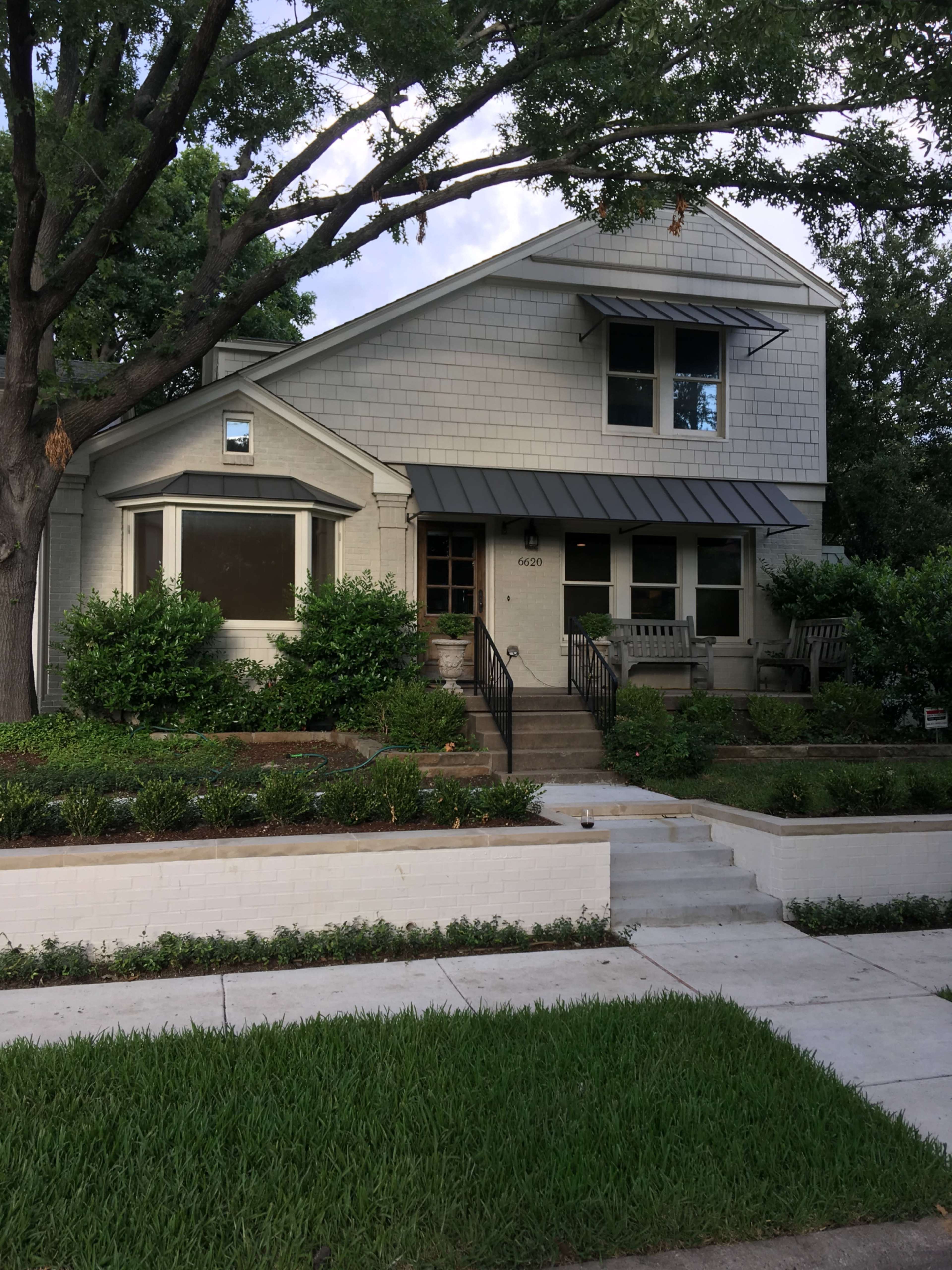 The image shows a two-story house with a gray shingle exterior, large front windows, and a landscaped yard with low greenery and a pathway.
