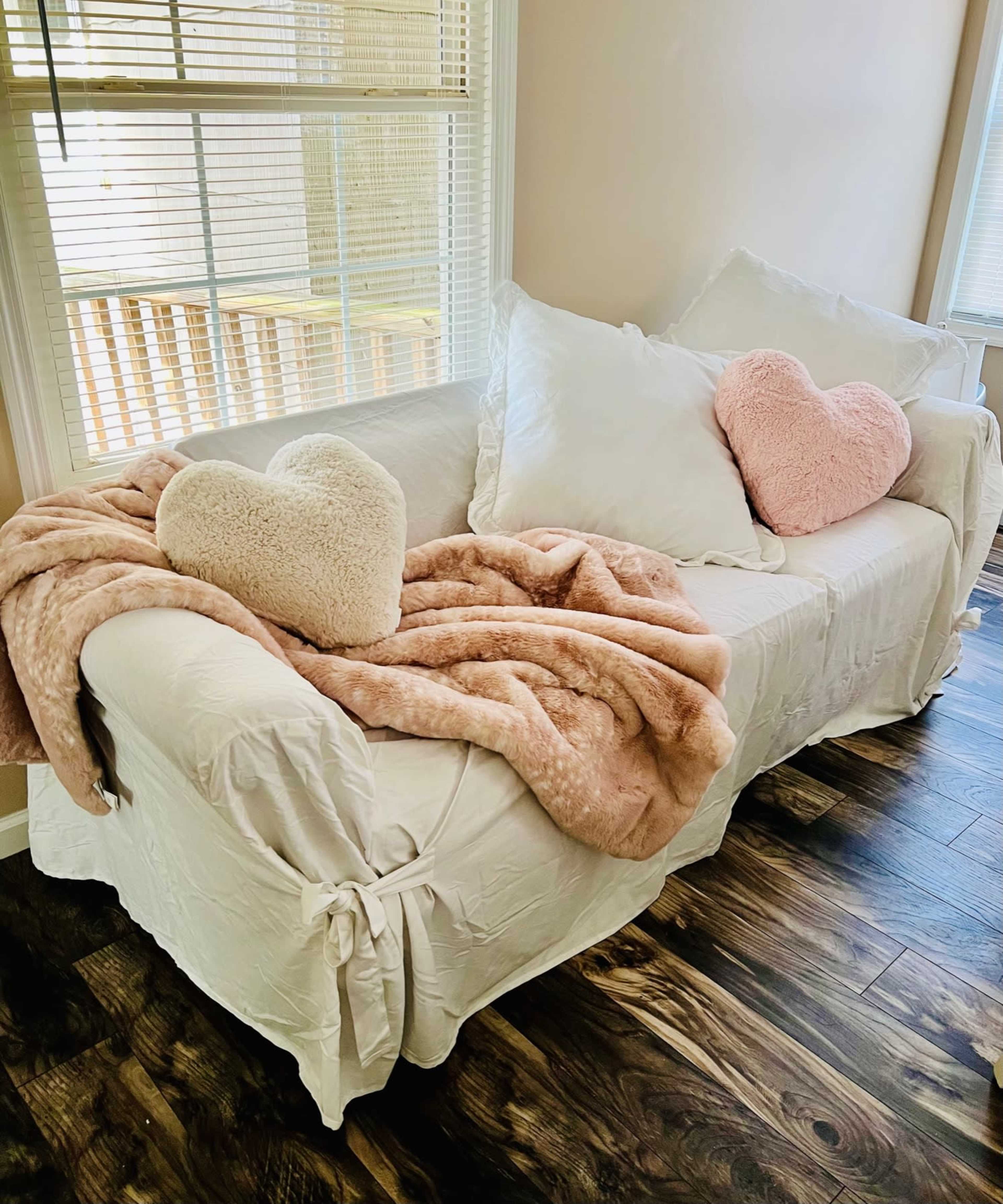 A couch with a white cover is adorned with a pink throw blanket and two heart-shaped pillows, one white and one pink, situated near a window.