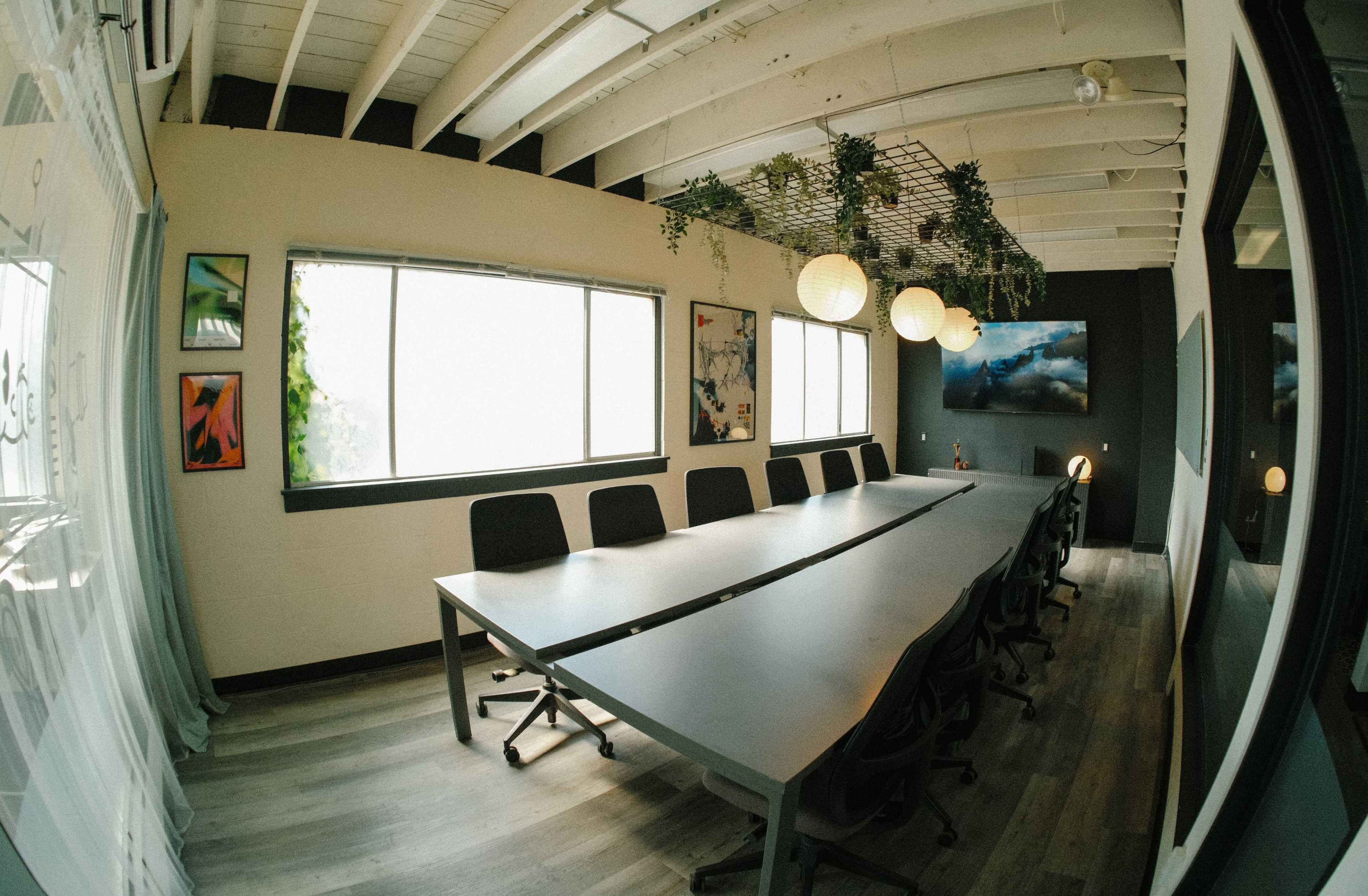 A modern conference room features a long table surrounded by chairs, with large windows and decorative plants overhead.