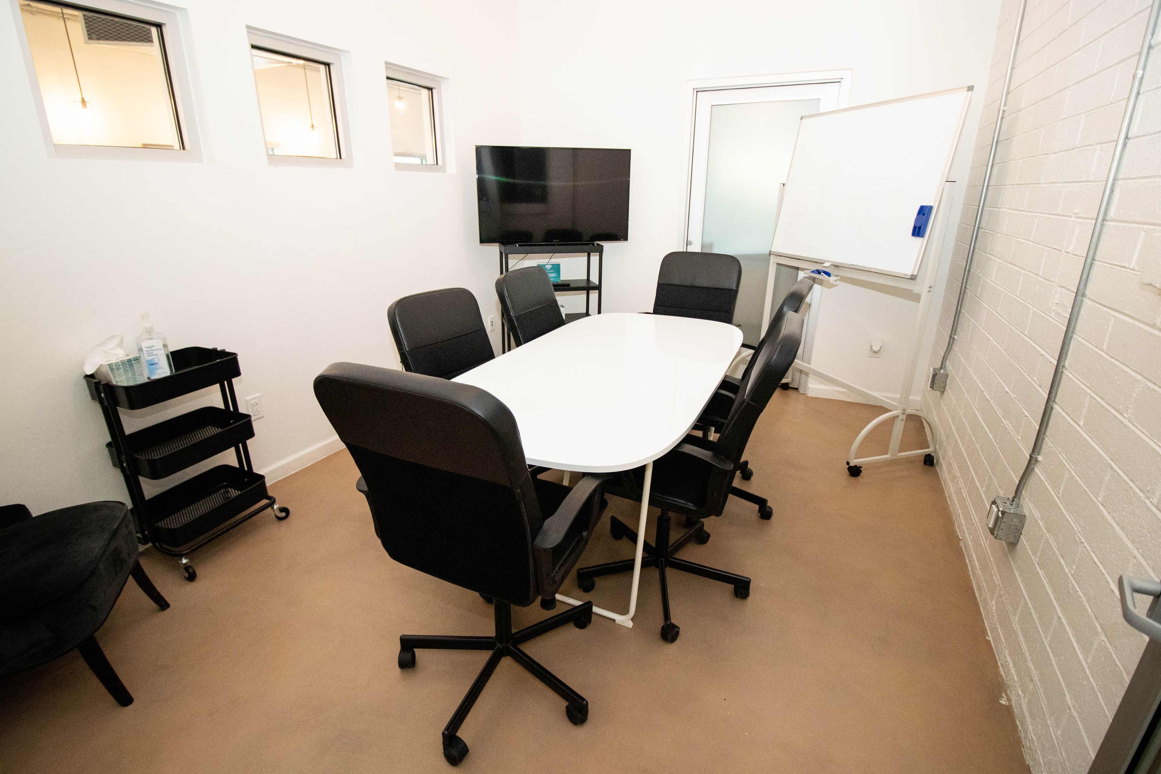 A small, well-lit conference room with a large white table surrounded by six black rolling chairs, a television on a stand, and a whiteboard on the wall.