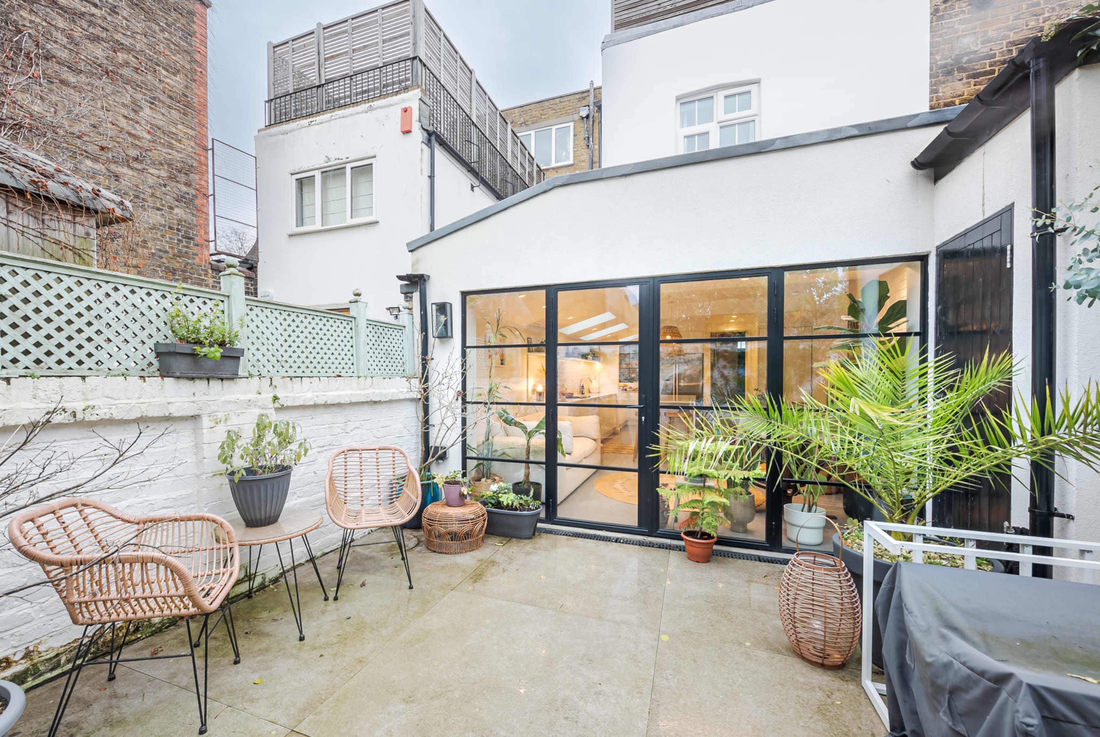 The image shows a small outdoor patio with two chairs and potted plants, adjacent to a house with large glass doors leading into a living space.