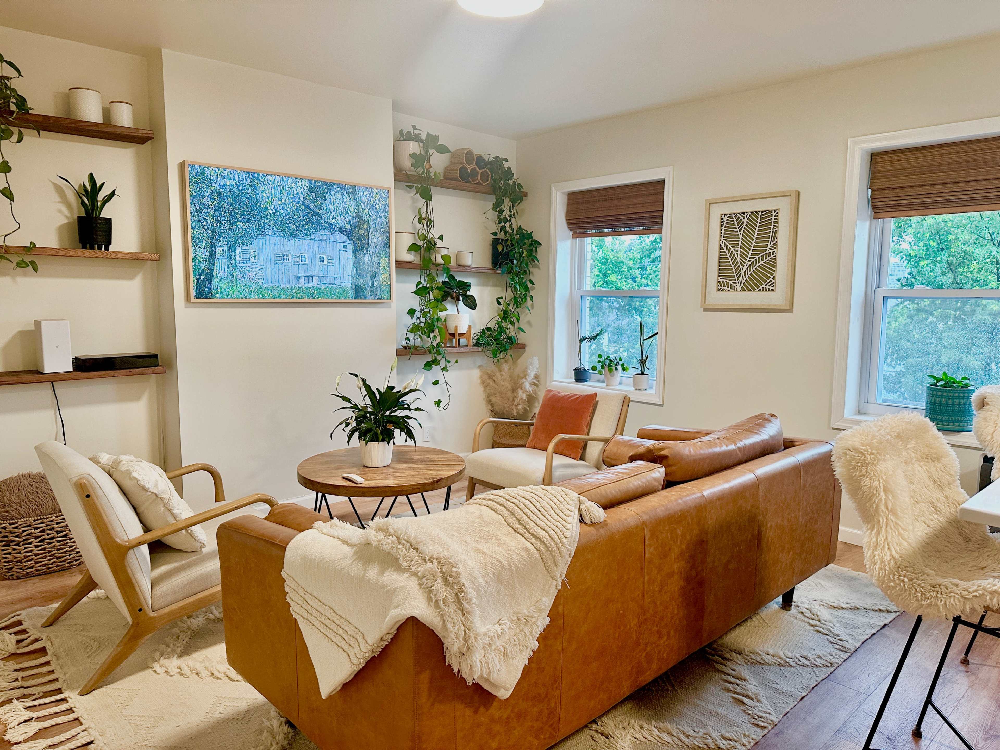 A cozy living room features a leather sofa, wooden shelves adorned with plants, and large windows allowing natural light to enter.